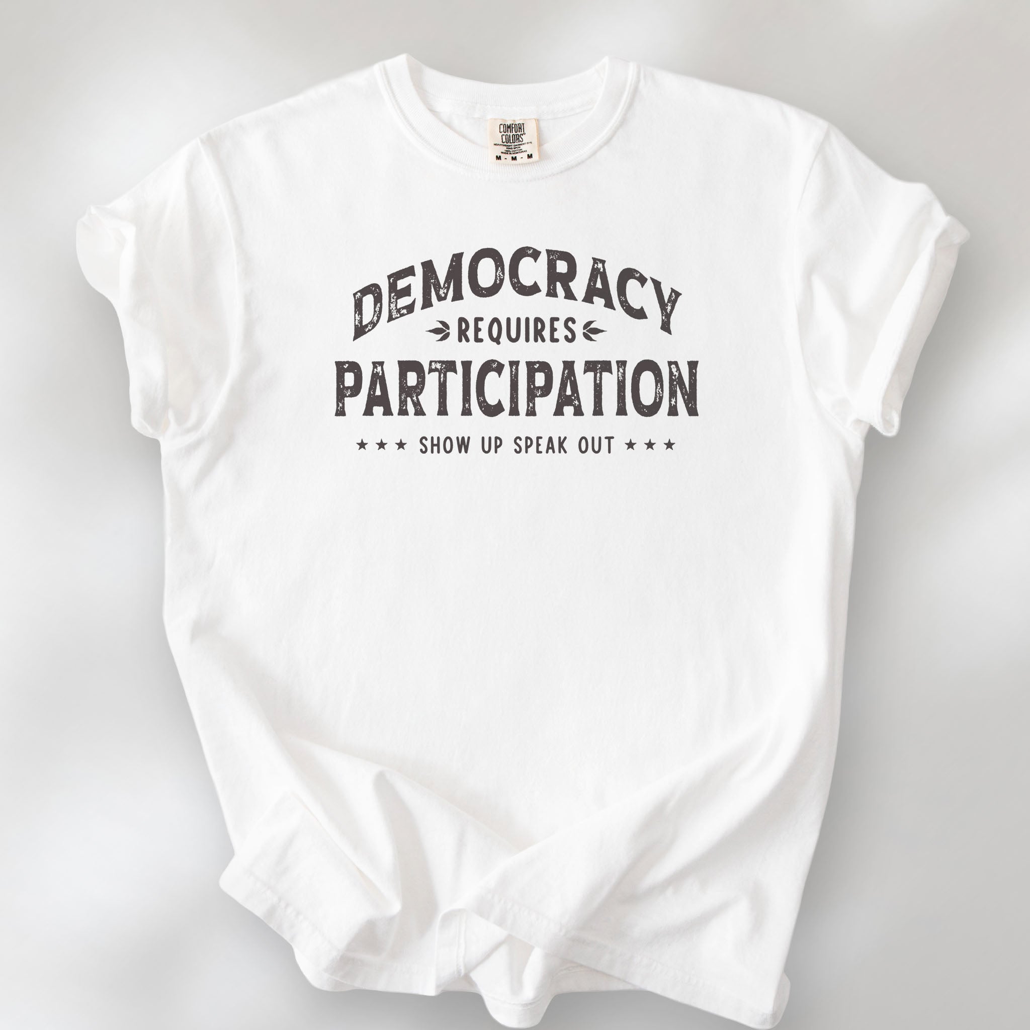 White t-shirt with vintage style typography reading “Democracy Requires Participation — Show Up Speak Out”.