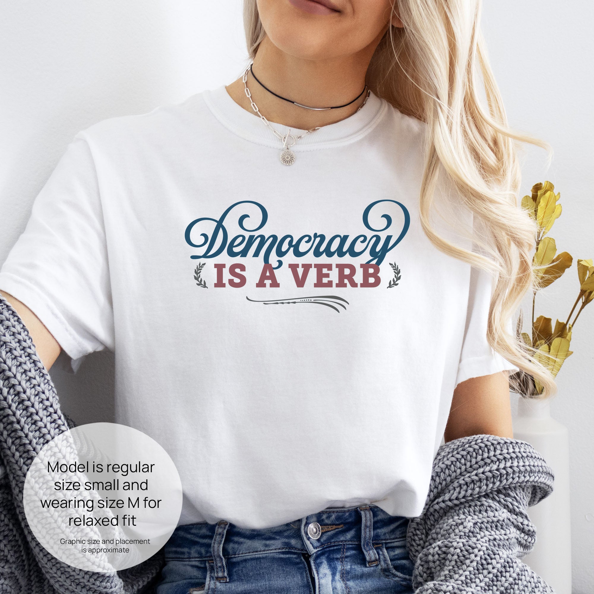 Female model wearing white t-shirt with the phrase “Democracy Is a Verb” in decorative lettering. Includes size and fit information for the model who is regular size small, and wearing a size M shirt for a relaxed fit.
