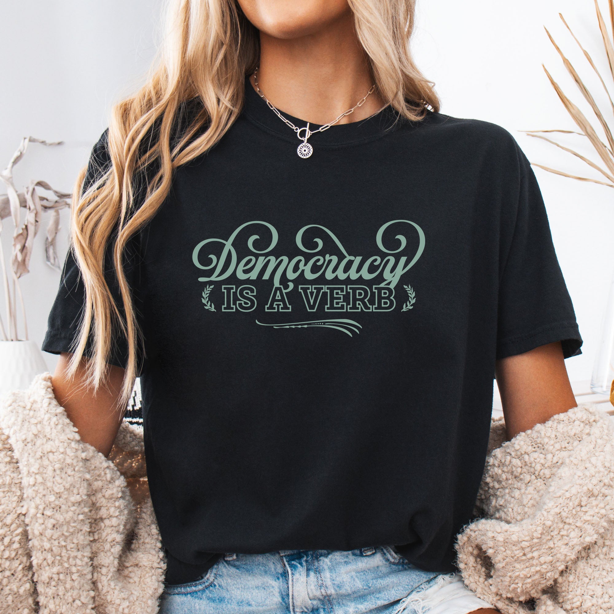 Female model wearing black t-shirt with the phrase “Democracy Is a Verb” in decorative mint green lettering.
