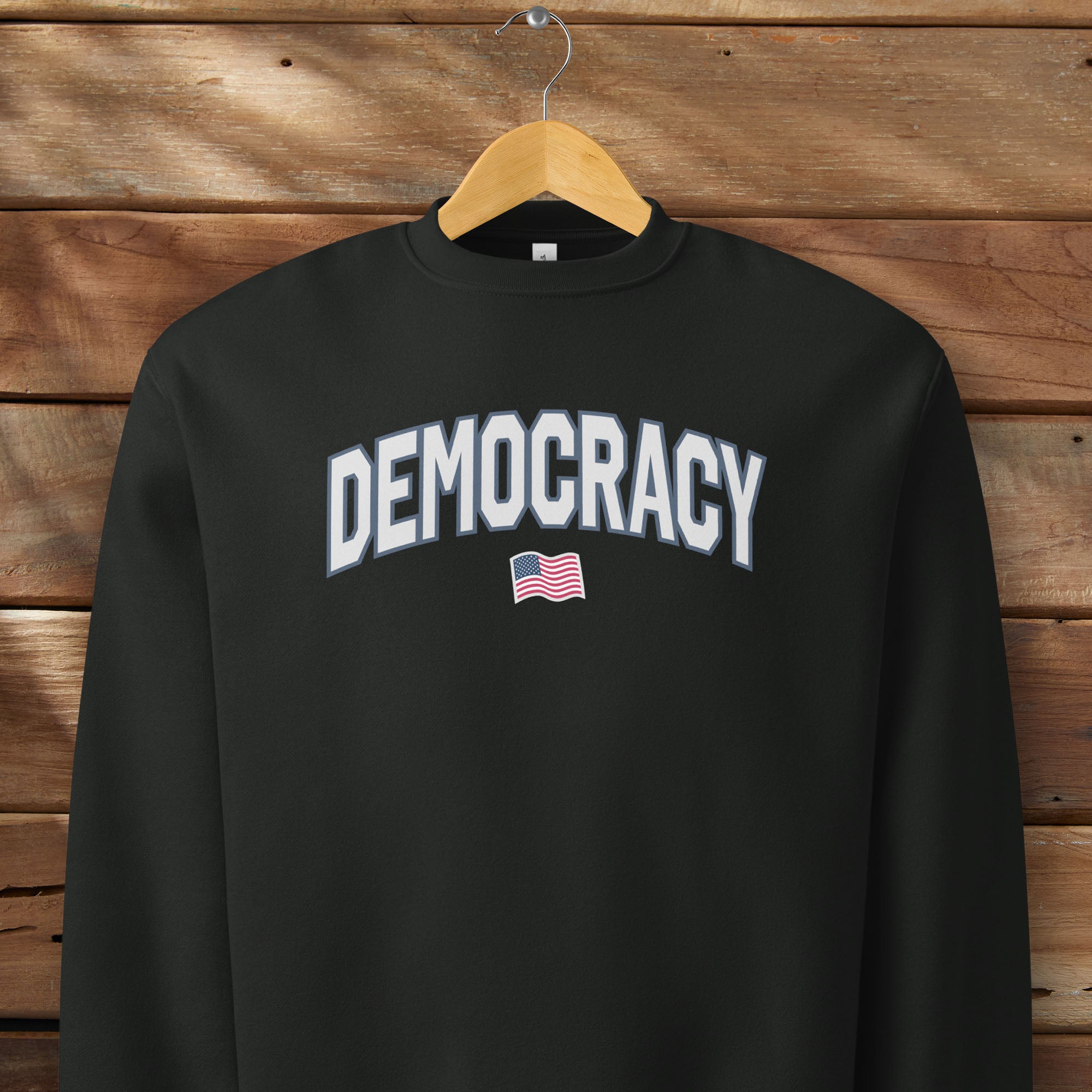 Black sweatshirt with the word “Democracy” in bold light grey letters and a small American flag underneath, symbolizing U.S. pride in democracy over authoritarianism, tyranny, dictators, handing on a hanger.