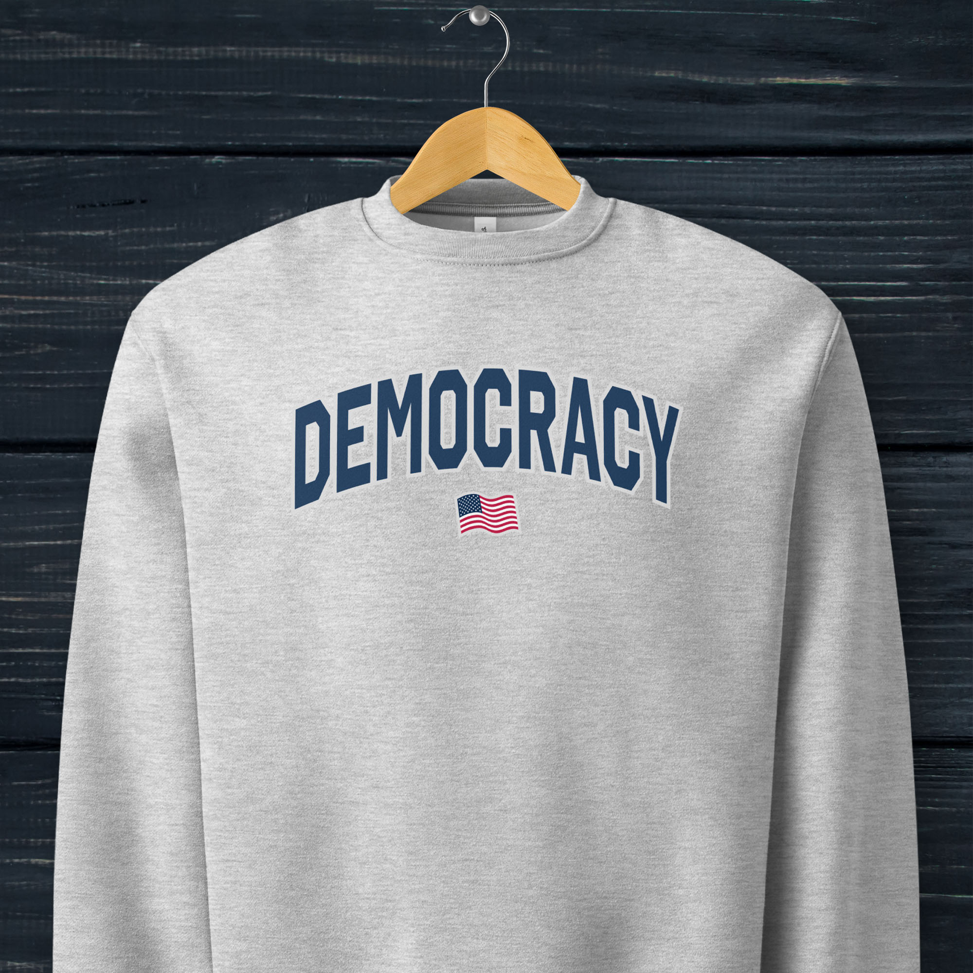 Athletic heather grey sweatshirt with the word “Democracy” in bold navy letters and a small American flag underneath, symbolizing U.S. pride in democracy over authoritarianism, tyranny, dictators, handing on a hanger.