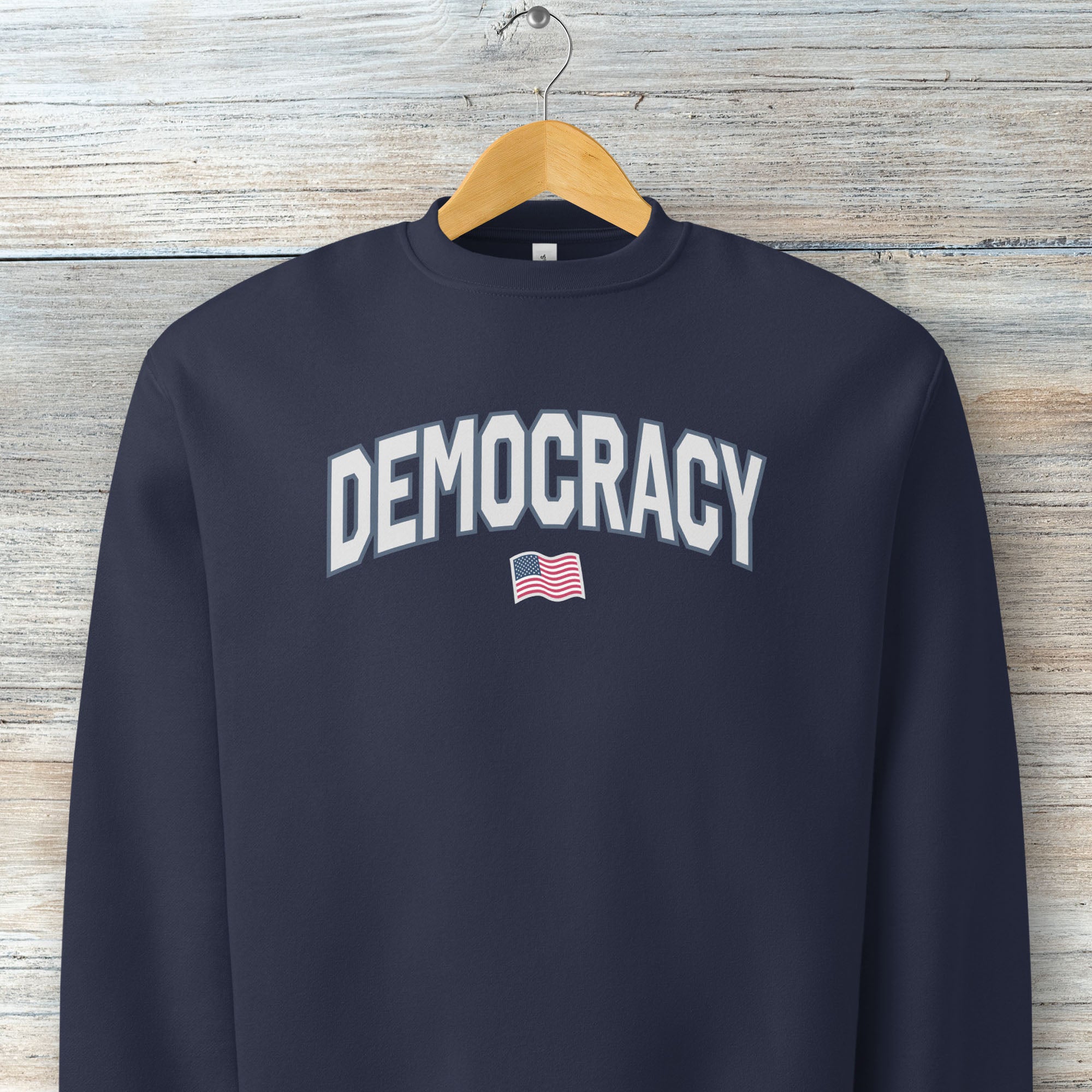 Navy sweatshirt with the word “Democracy” in bold light grey letters and a small American flag underneath, symbolizing U.S. pride in democracy over authoritarianism, tyranny, dictators, handing on a hanger.