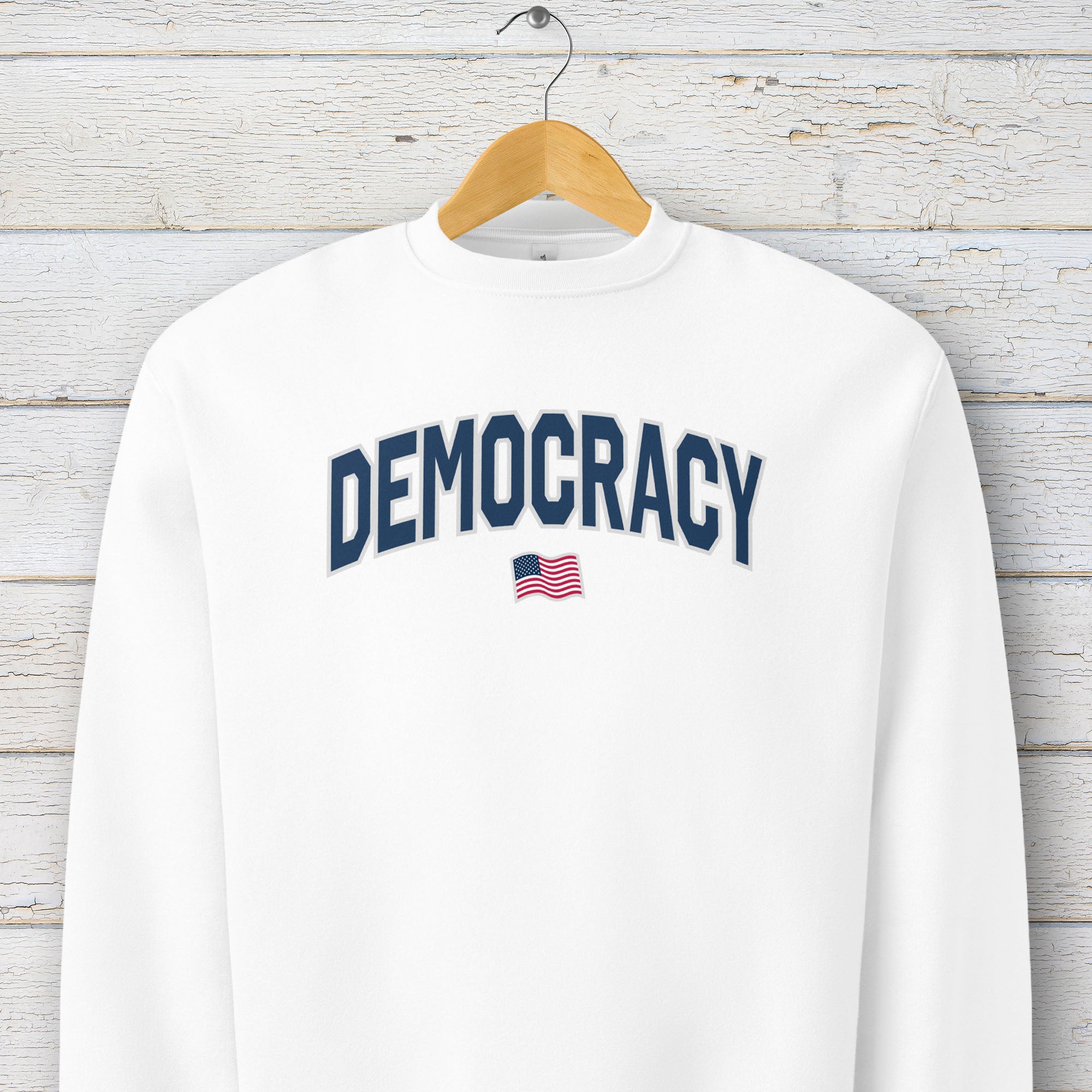 White sweatshirt with the word “Democracy” in bold navy letters and a small American flag underneath, symbolizing U.S. pride in democracy over authoritarianism, tyranny, dictators, handing on a hanger.