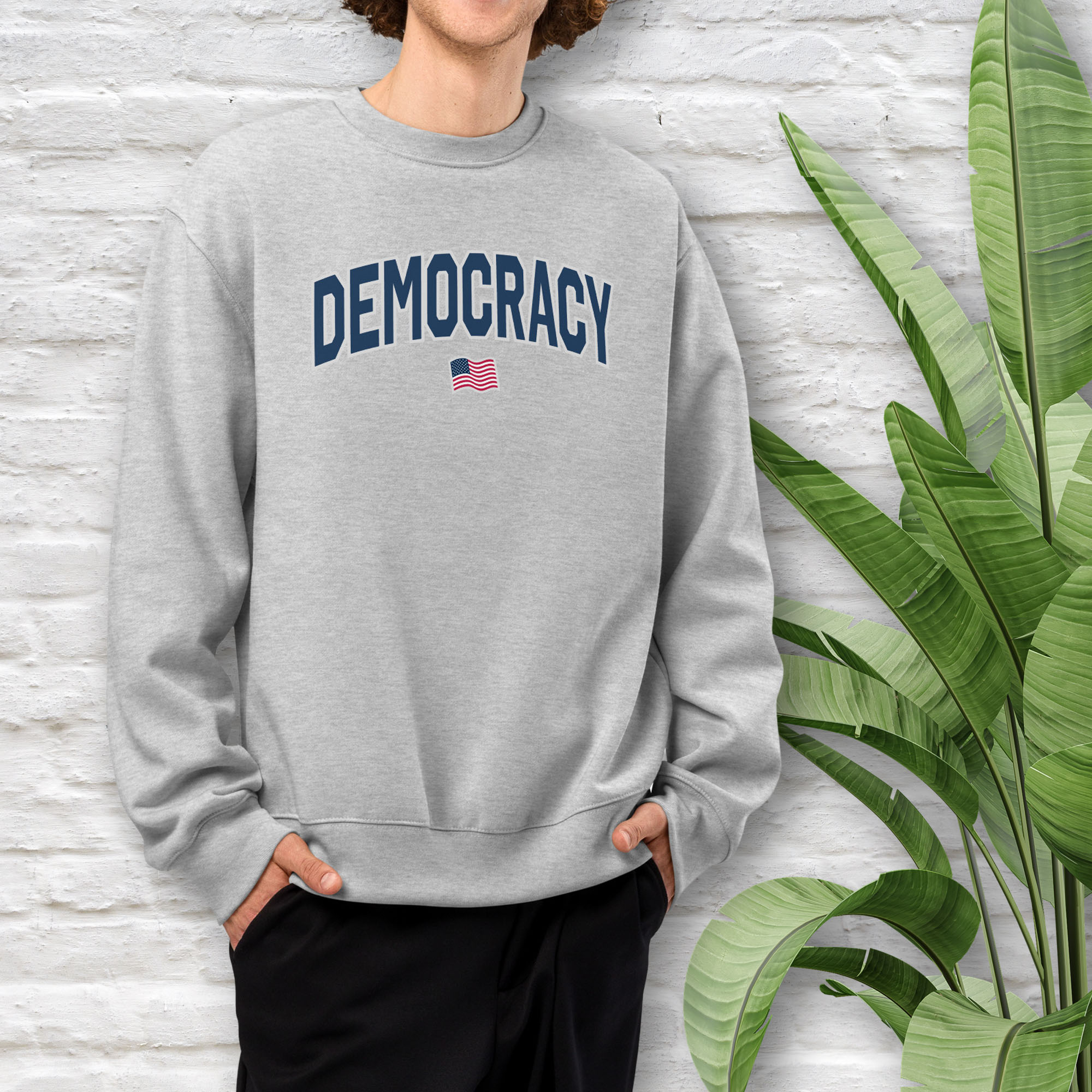 Male model wearing athletic heather grey sweatshirt with the word “Democracy” in bold navy letters and a small American flag underneath, symbolizing U.S. pride in democracy over authoritarianism, tyranny, dictators.