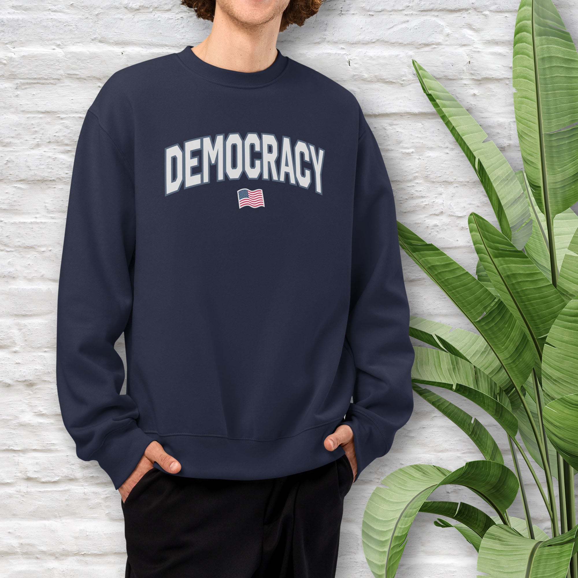 Male model wearing navy sweatshirt with the word “Democracy” in bold light grey letters and a small American flag underneath, symbolizing U.S. pride in democracy over authoritarianism, tyranny, dictators.