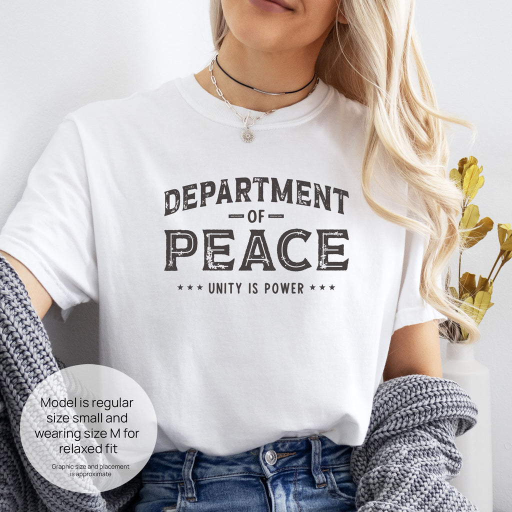 Female model wearing white t-shirt with distressed text reading “Department of Peace — Unity is Power” in vintage style design. Includes size and fit information for the model who is regular size small, and wearing a size M shirt for a relaxed fit.
