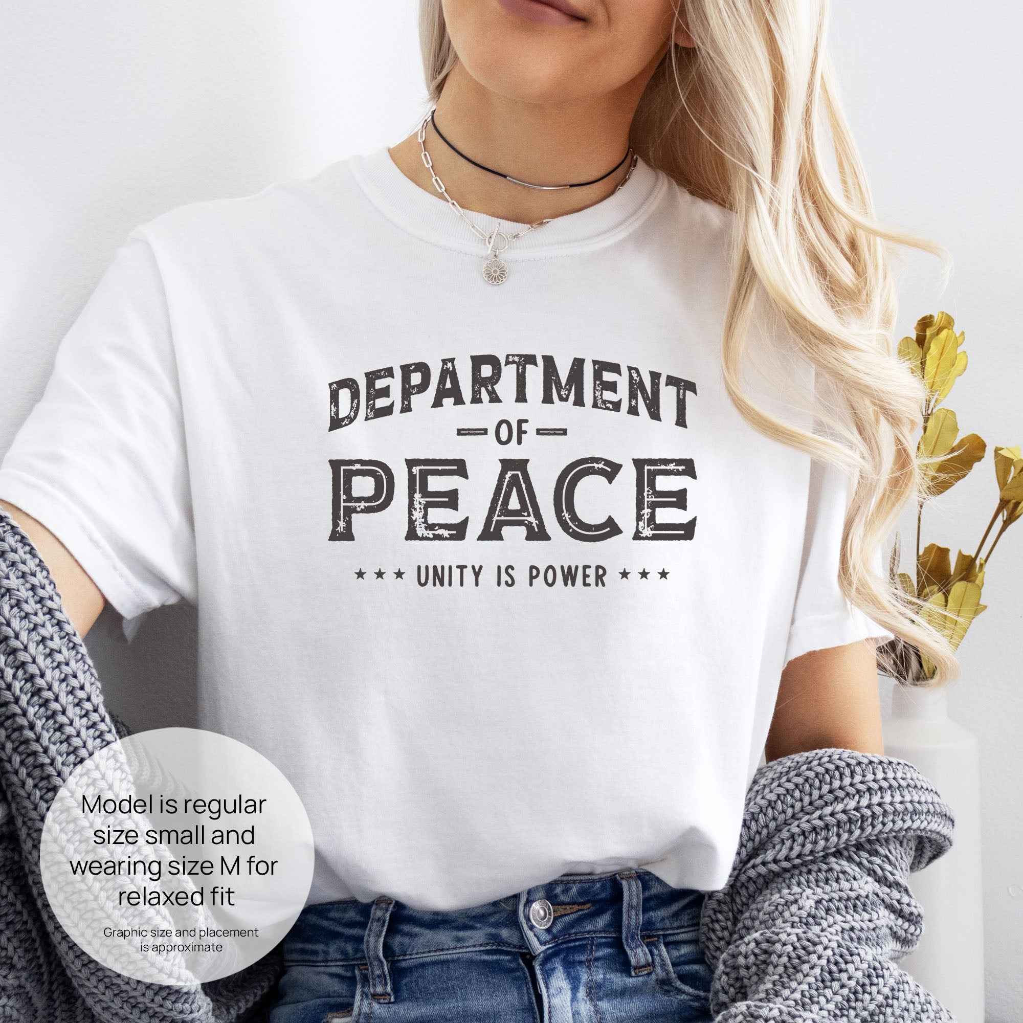 Female model wearing white t-shirt with distressed text reading “Department of Peace — Unity is Power” in vintage style design. Includes size and fit information for the model who is regular size small, and wearing a size M shirt for a relaxed fit.
