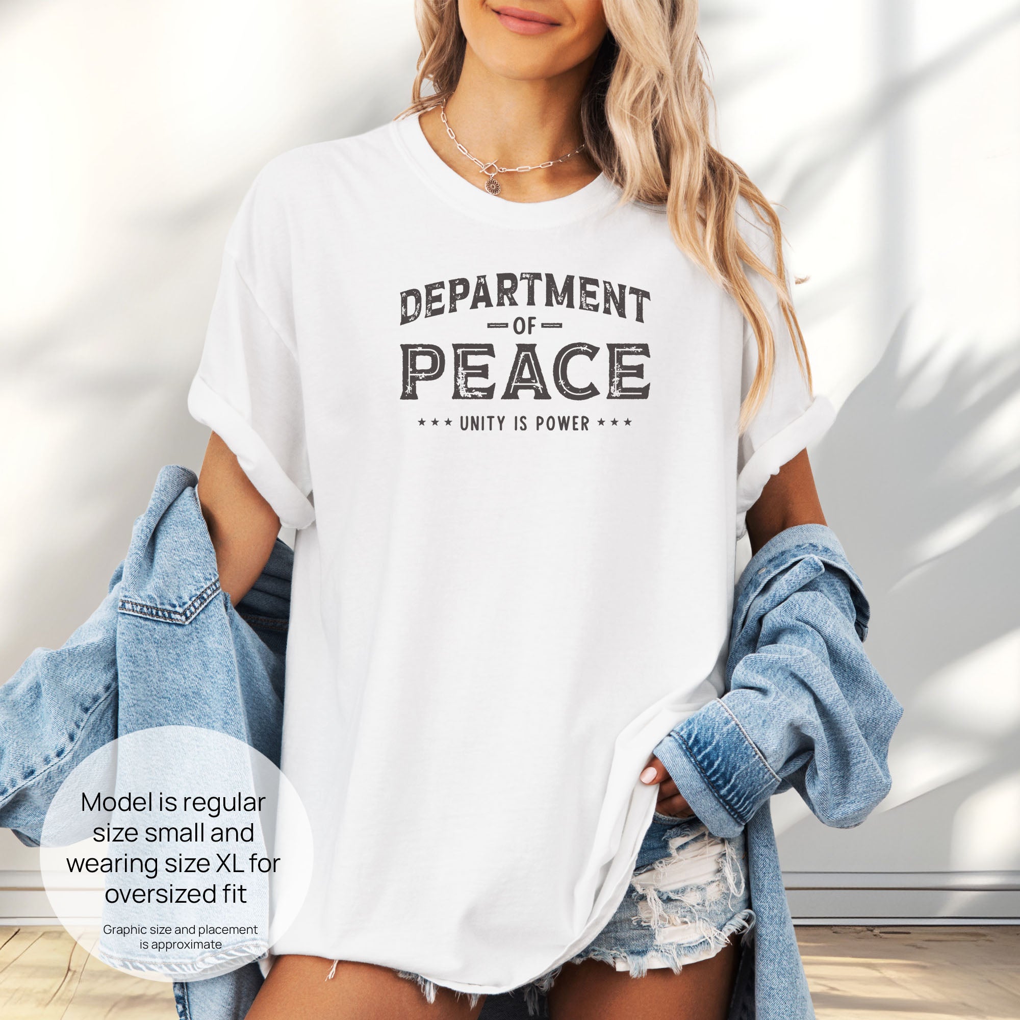 Female model wearing oversized white t-shirt with distressed text reading “Department of Peace — Unity is Power” in vintage style design. Includes size and fit information for the model who is regular size small, and wearing a size XL shirt for an oversized fit.
