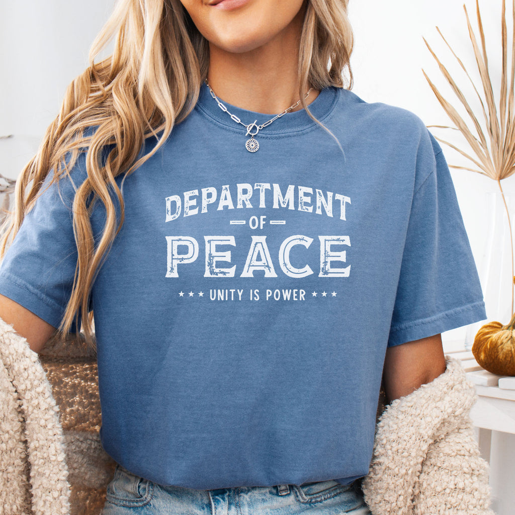 Female model wearing blue jean t-shirt with distressed text reading “Department of Peace — Unity is Power” in vintage style design.