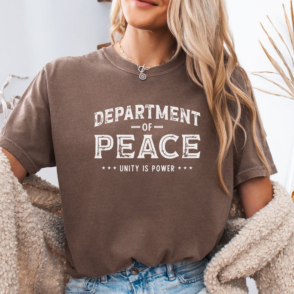 Female model wearing espresso t-shirt with distressed text reading “Department of Peace — Unity is Power” in vintage style design.