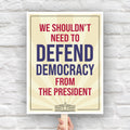 Downloadable, printable protest sign with a light gold background and red and blue text that reads "we shouldn't need to defend democracy from the president".