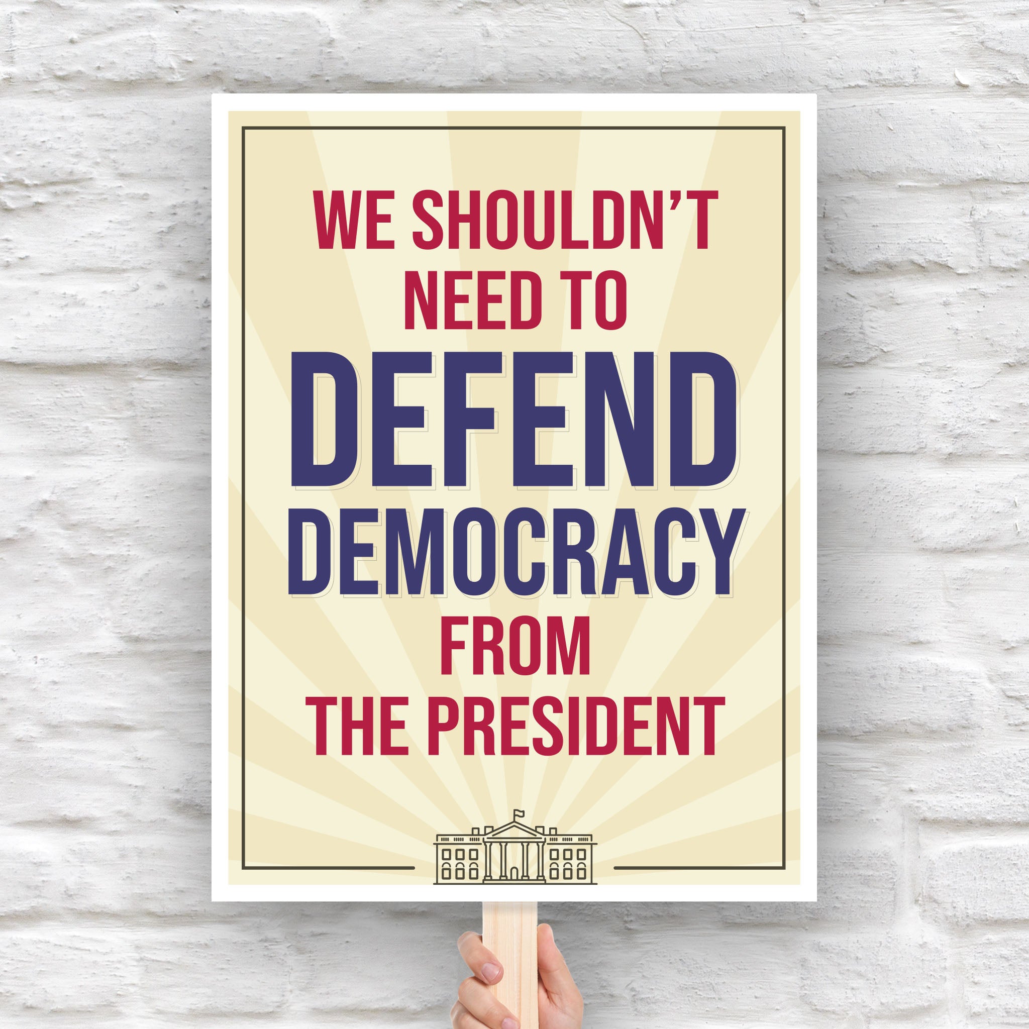 Downloadable, printable protest sign with a light gold background and red and blue text that reads "we shouldn't need to defend democracy from the president".
