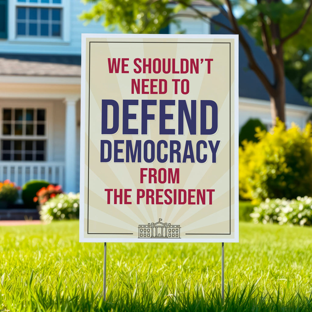 Yard sign in a green lawn, with a light gold background and red and blue text that reads "we shouldn't need to defend democracy from the president".
