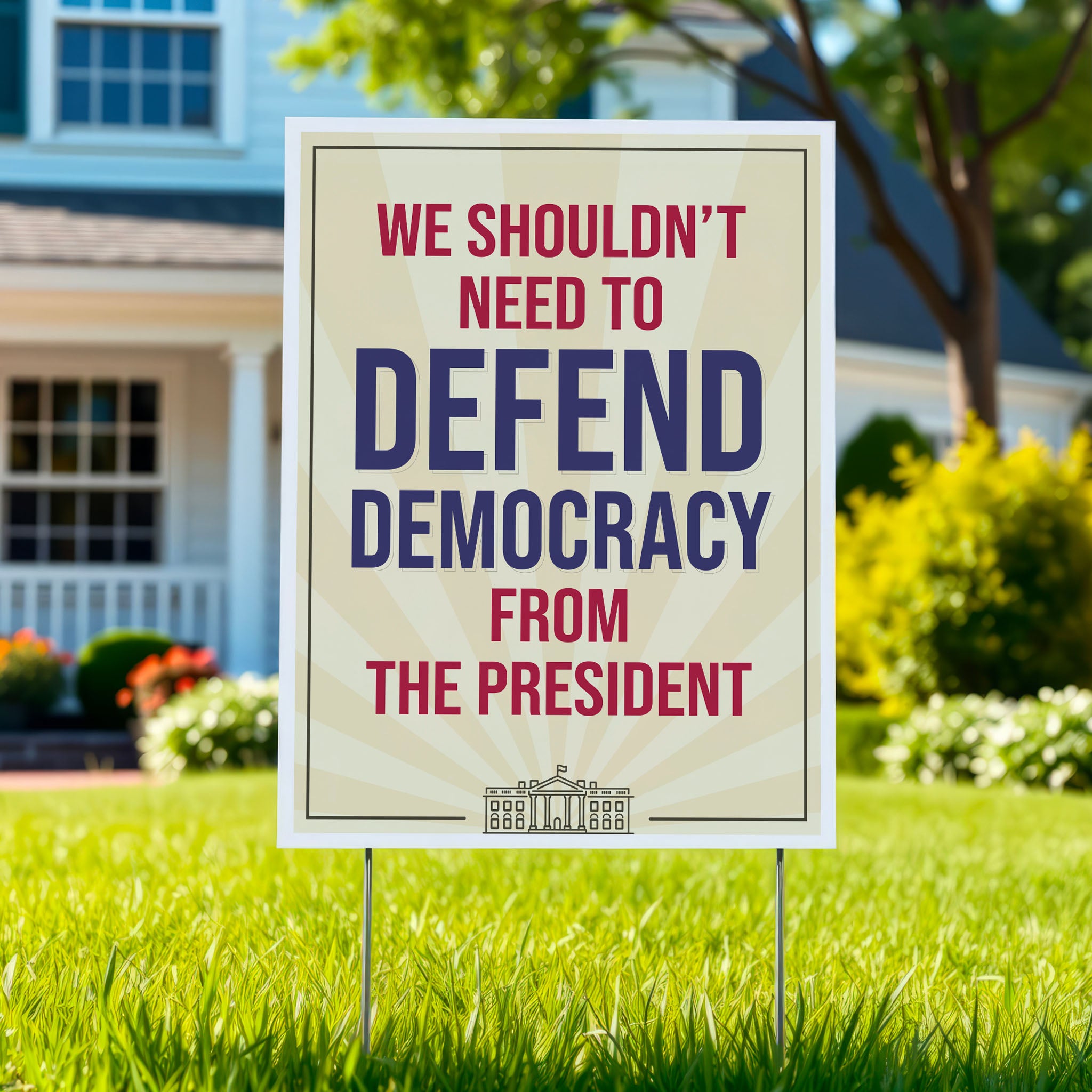 Yard sign in a green lawn, with a light gold background and red and blue text that reads "we shouldn't need to defend democracy from the president".