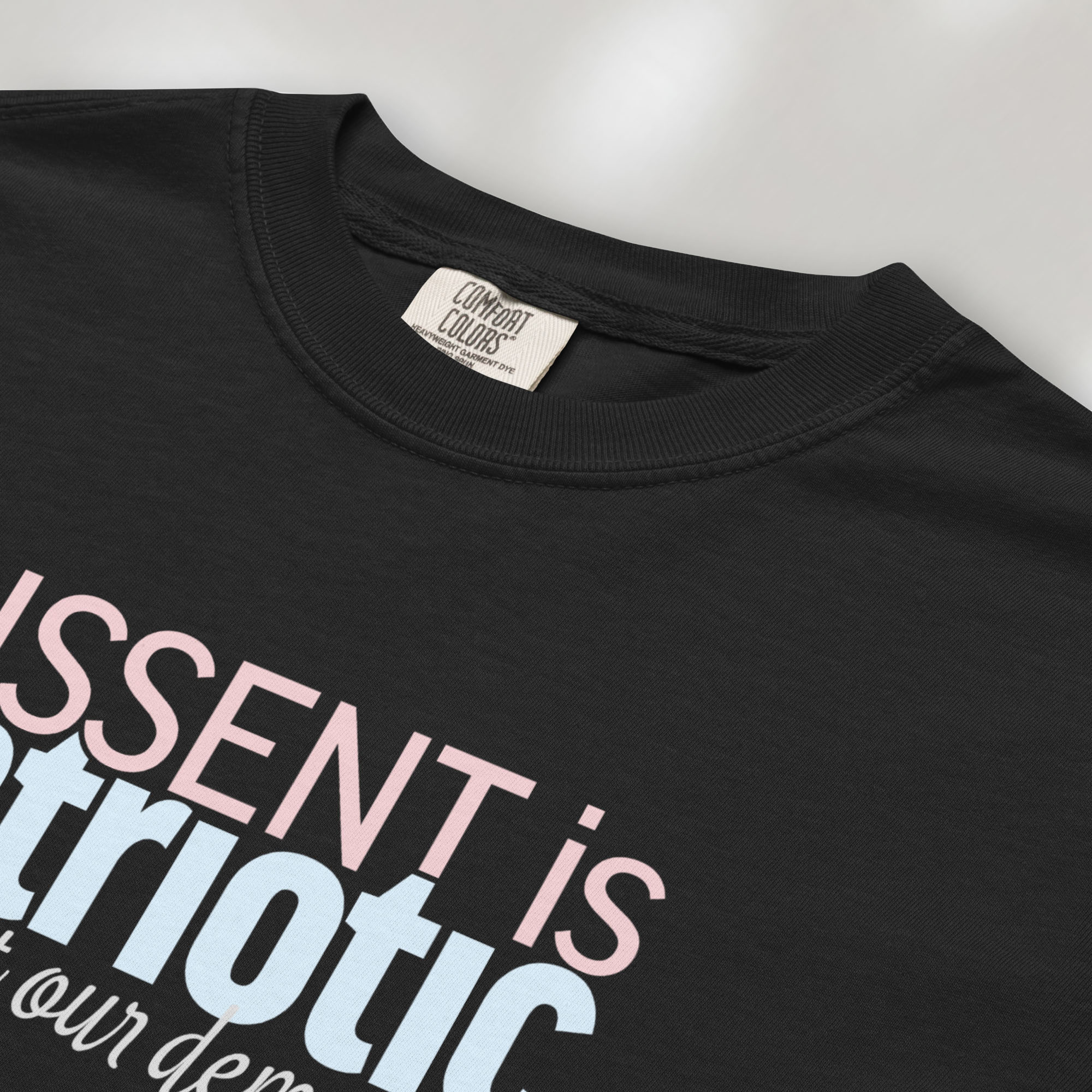 Closeup detail of black t-shirt with bold stylized red and blue text that reads "dissent is patriotic" with script text below that reads "protest our democracy".
