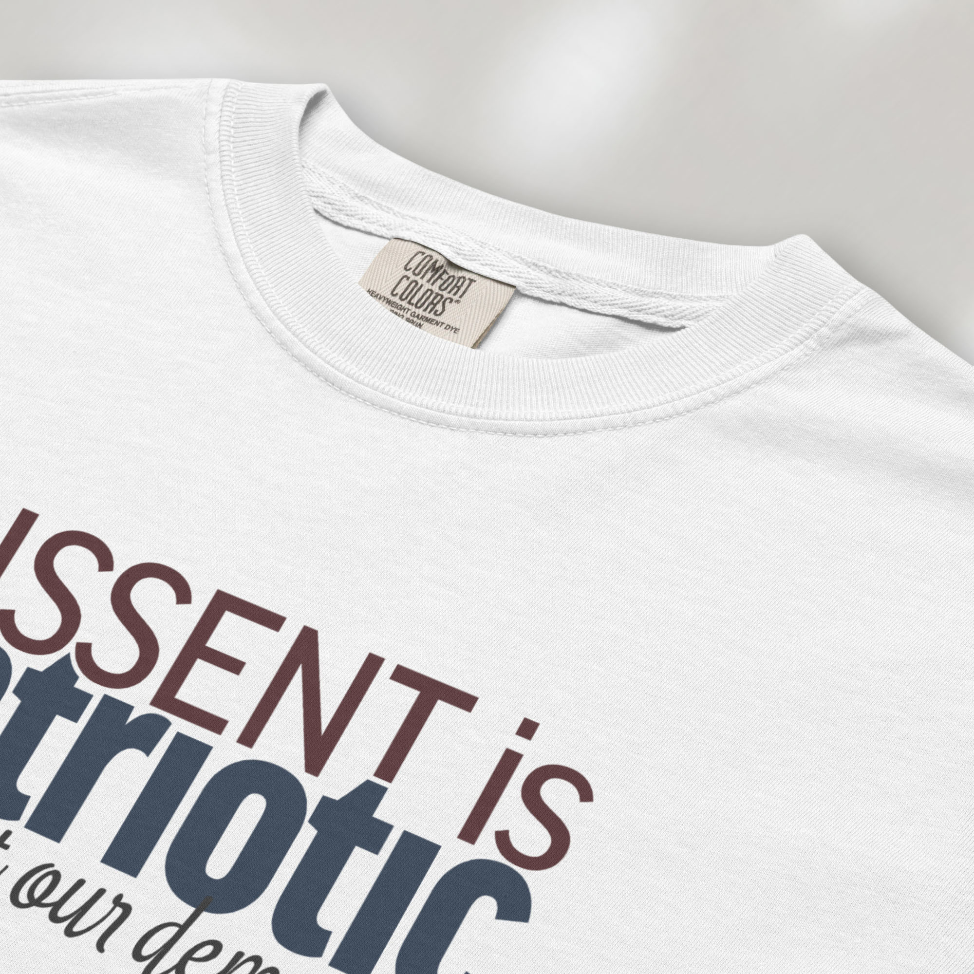 Closeup detail of white t-shirt with bold stylized red and blue text that reads "dissent is patriotic" with script text below that reads "protest our democracy".