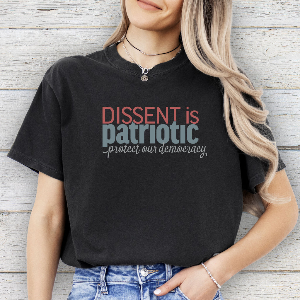 Female model wearing black t-shirt with bold stylized red and blue text that reads "dissent is patriotic" with script text below that reads "protest our democracy".