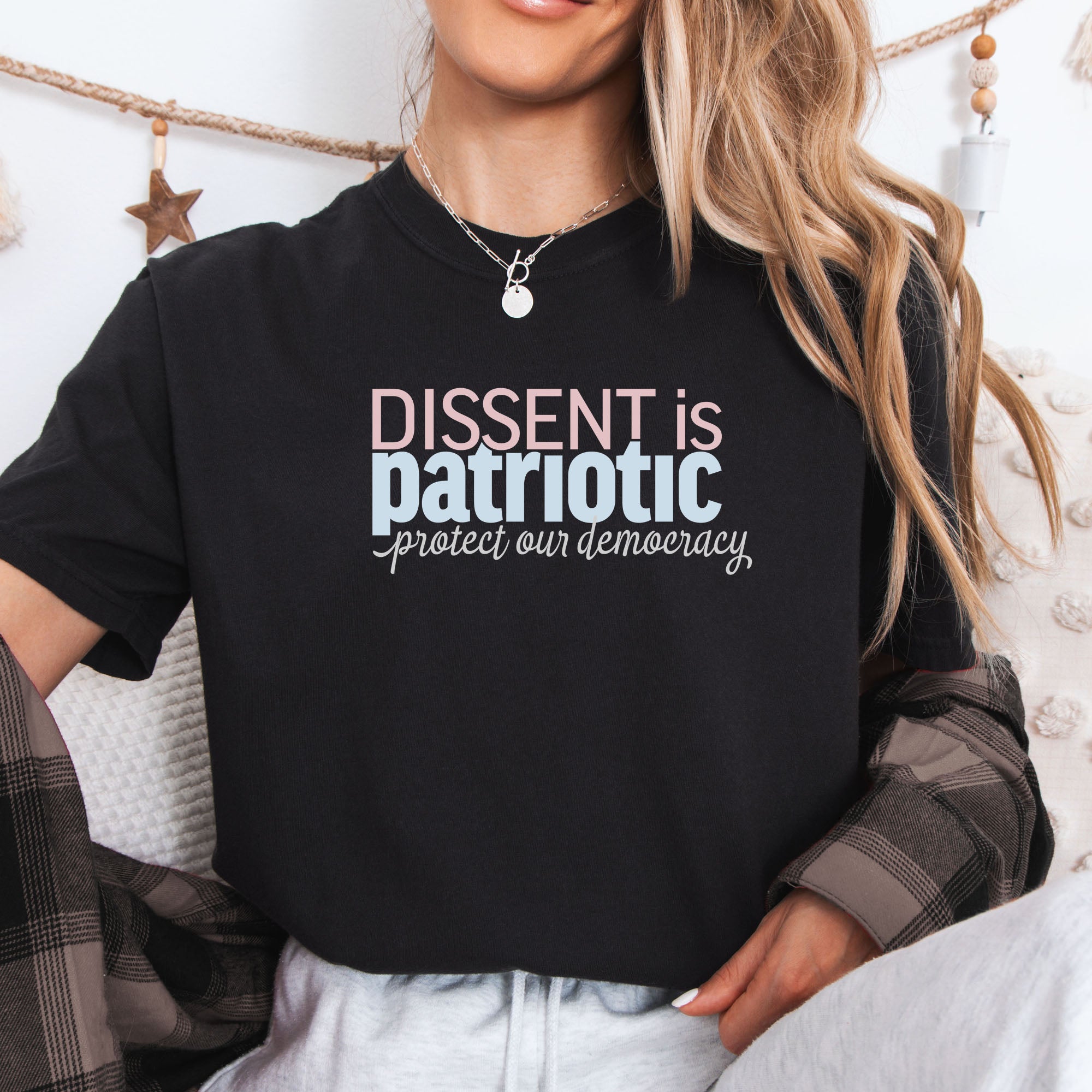 Female model wearing black t-shirt with bold stylized red and blue text that reads "dissent is patriotic" with script text below that reads "protest our democracy".
