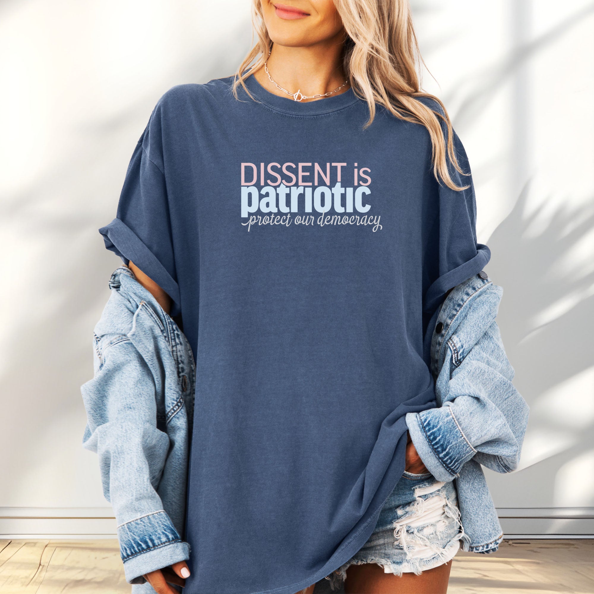 Female model wearing oversized denim t-shirt with bold stylized red and blue text that reads "dissent is patriotic" with script text below that reads "protest our democracy".