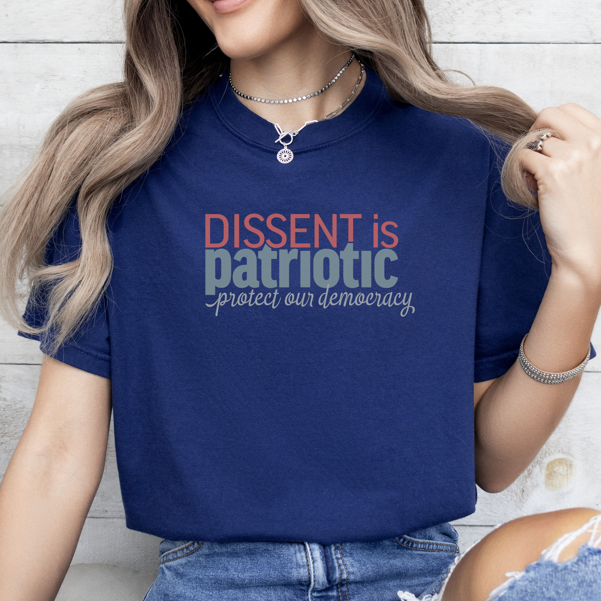 Female model wearing true navy t-shirt with bold stylized red and blue text that reads "dissent is patriotic" with script text below that reads "protest our democracy".