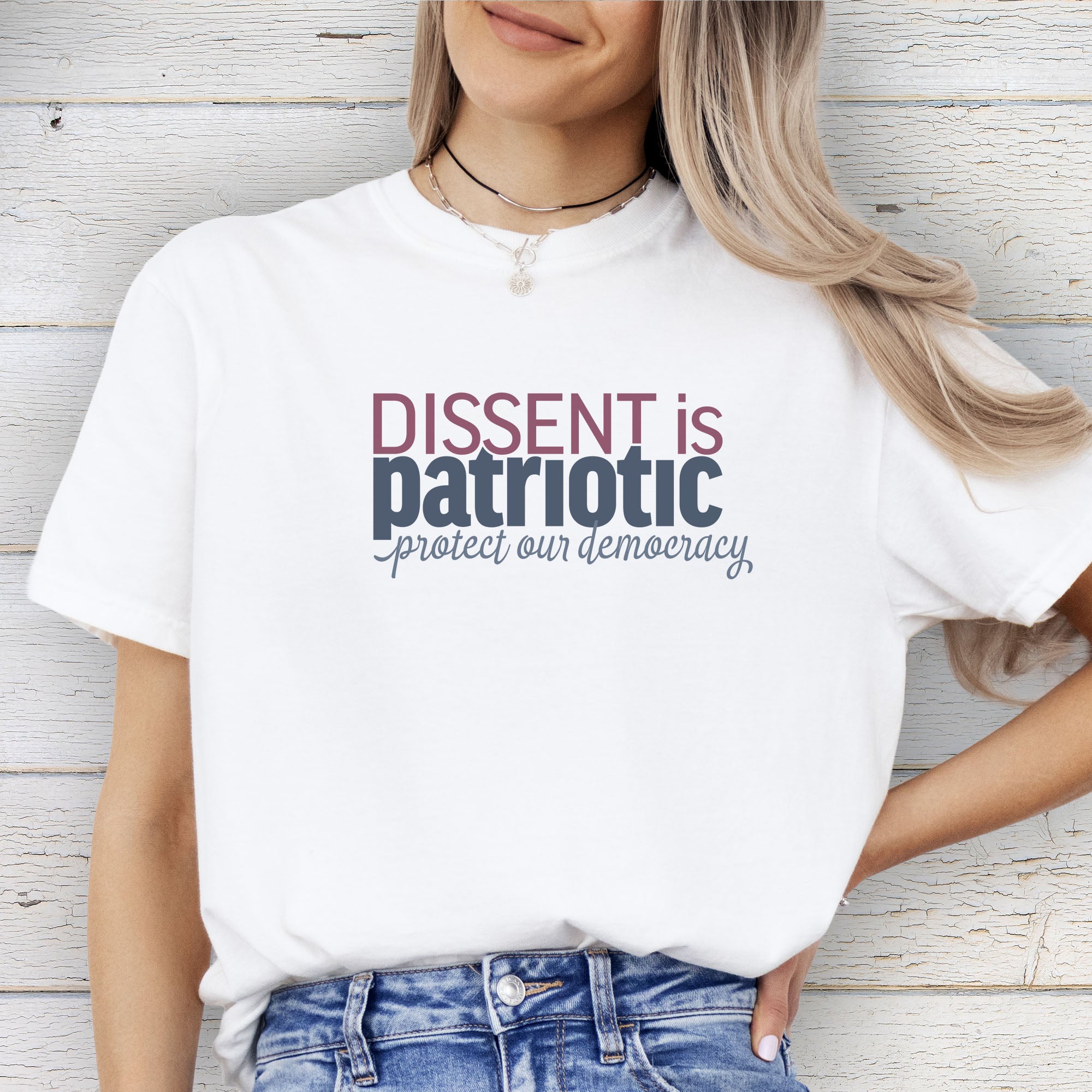 Female model wearing a white unisex t-shirt with bold text reading “Dissent is Patriotic – Protect Our Democracy” in red, blue, and gray.