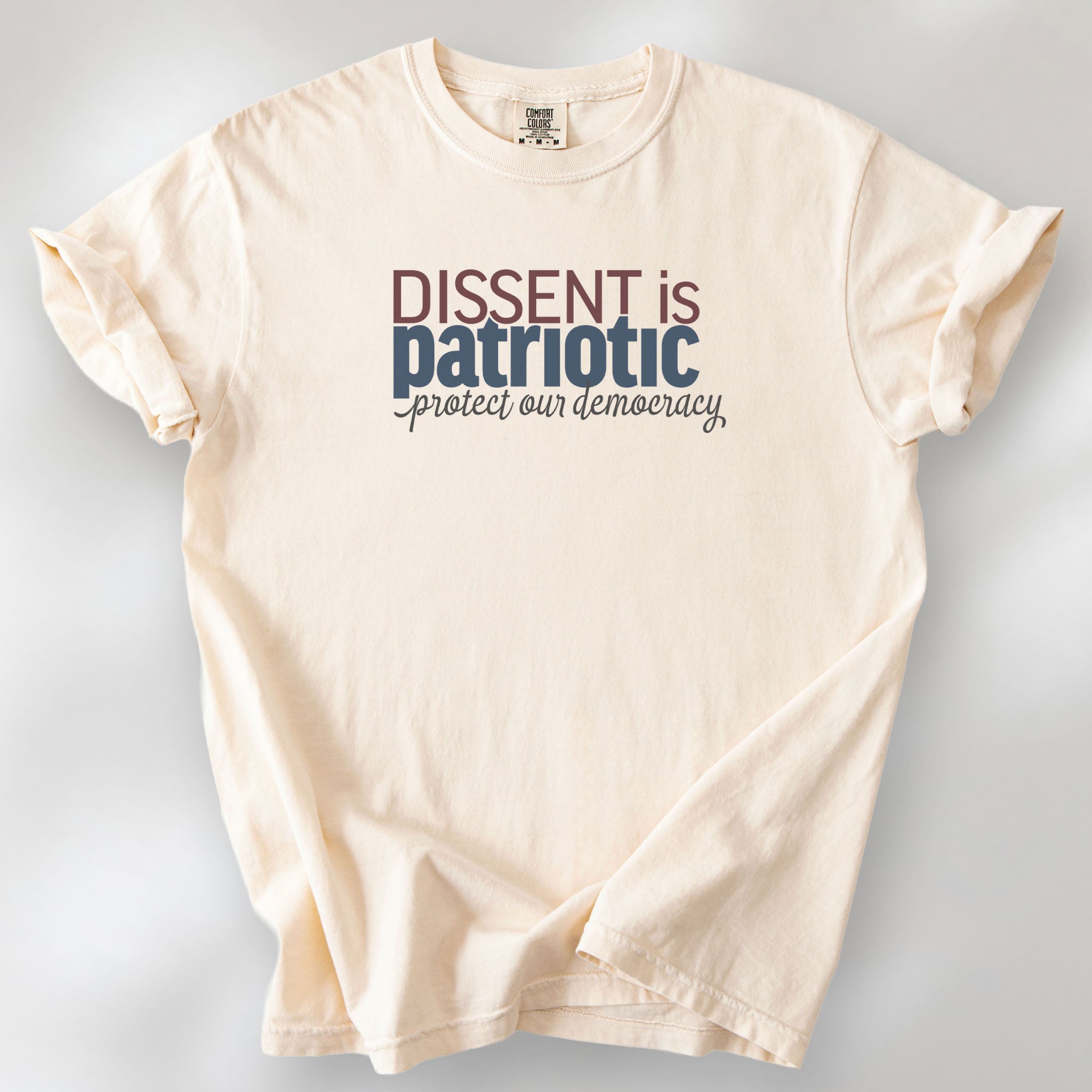 Ivory t-shirt with bold stylized red and blue text that reads "dissent is patriotic" with script text below that reads "protest our democracy".