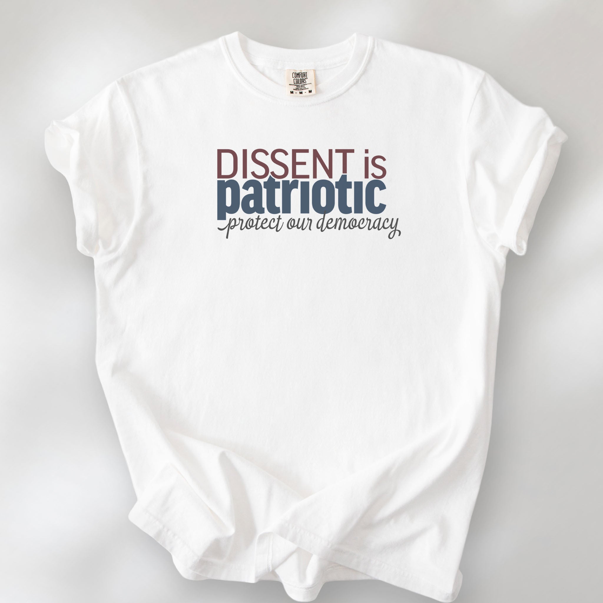 White t-shirt with bold stylized red and blue text that reads "dissent is patriotic" with script text below that reads "protest our democracy".