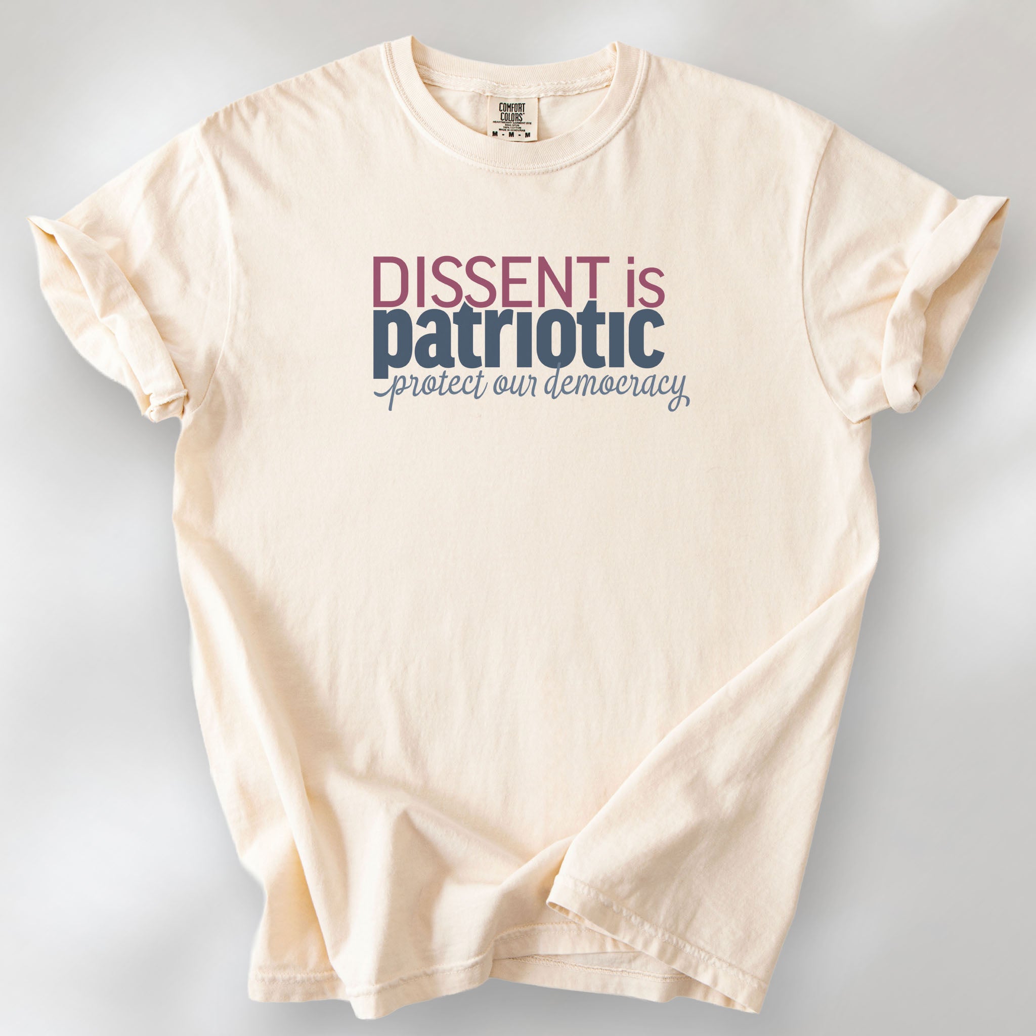 Ivory unisex t-shirt with bold text reading “Dissent is Patriotic – Protect Our Democracy” in red, blue, and gray.