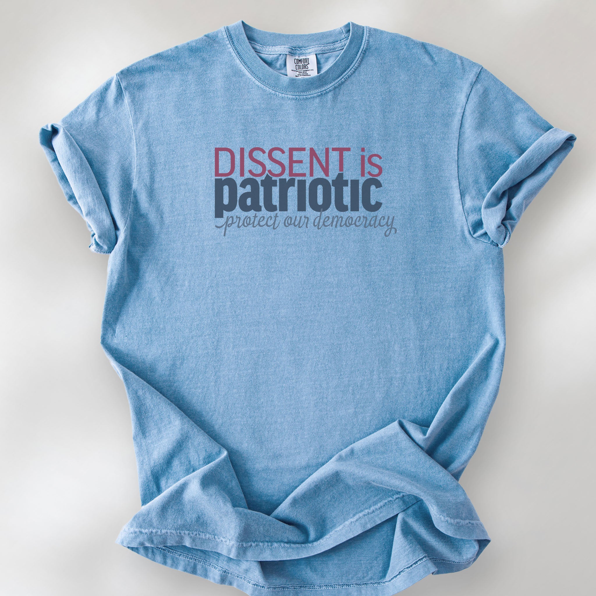 Washed denim unisex t-shirt with bold text reading “Dissent is Patriotic – Protect Our Democracy” in red, blue, and gray.