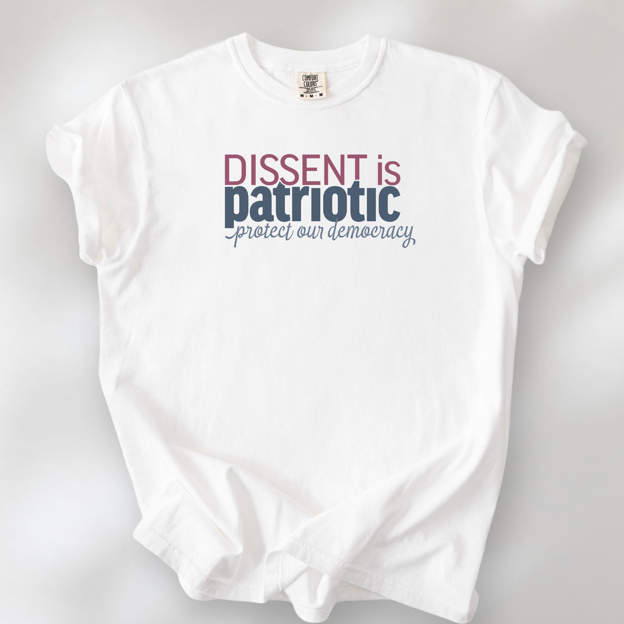 White unisex t-shirt with bold text reading “Dissent is Patriotic – Protect Our Democracy” in red, blue, and gray.