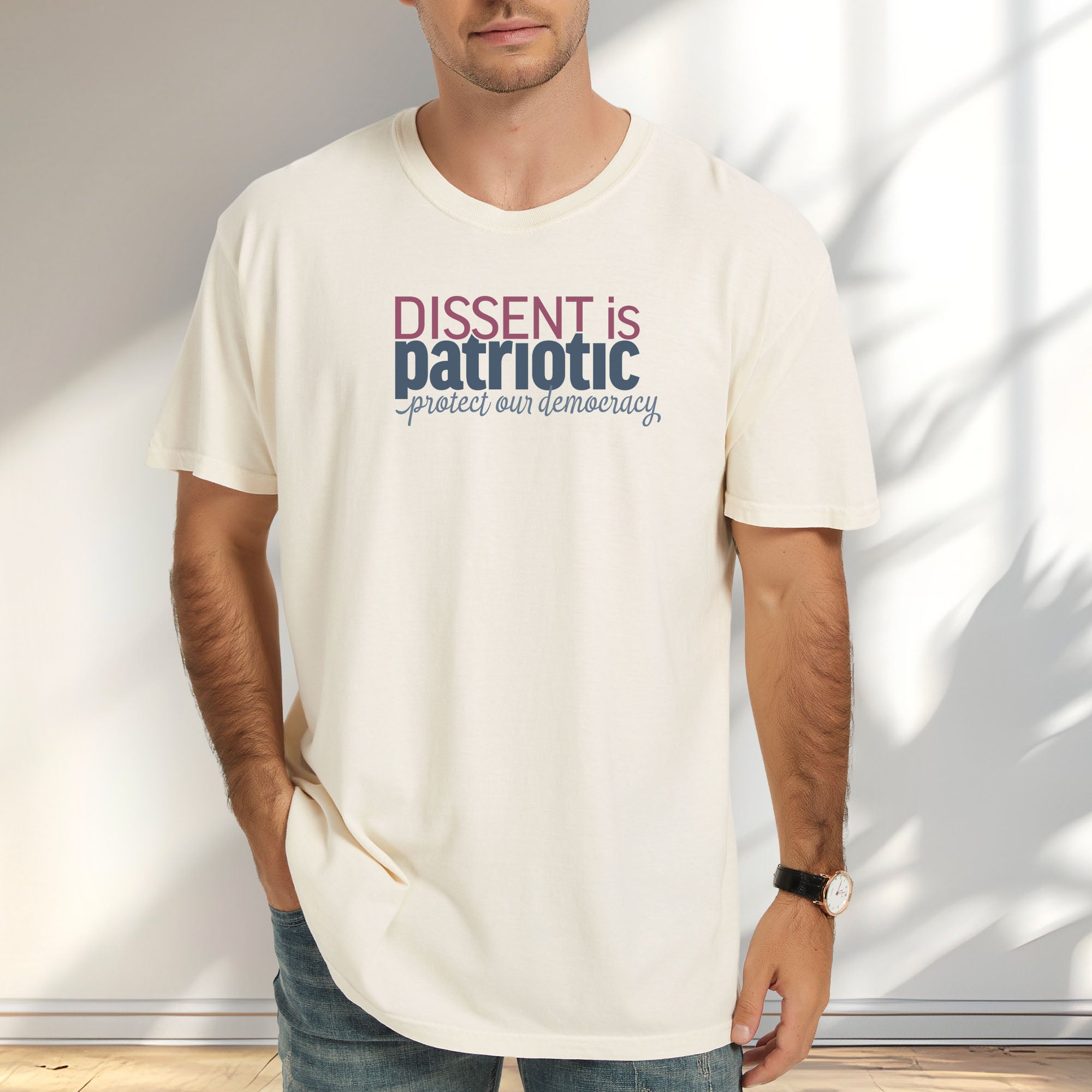 Male model wearing ivory unisex t-shirt with bold text reading “Dissent is Patriotic – Protect Our Democracy” in red, blue, and gray.