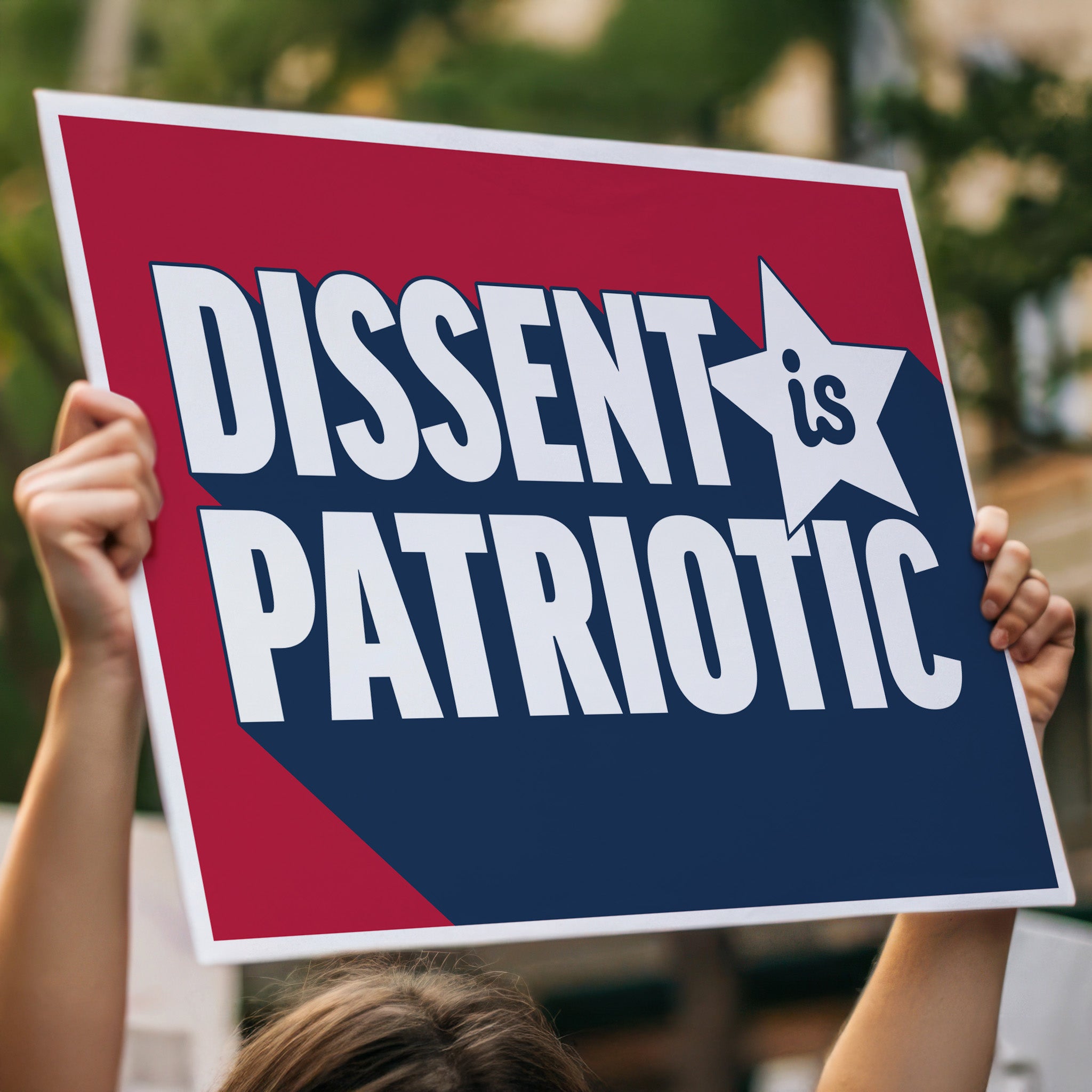 Person holding red, white and blue protest sign with bold text that reads “Dissent is Patriotic” high in a crowd.