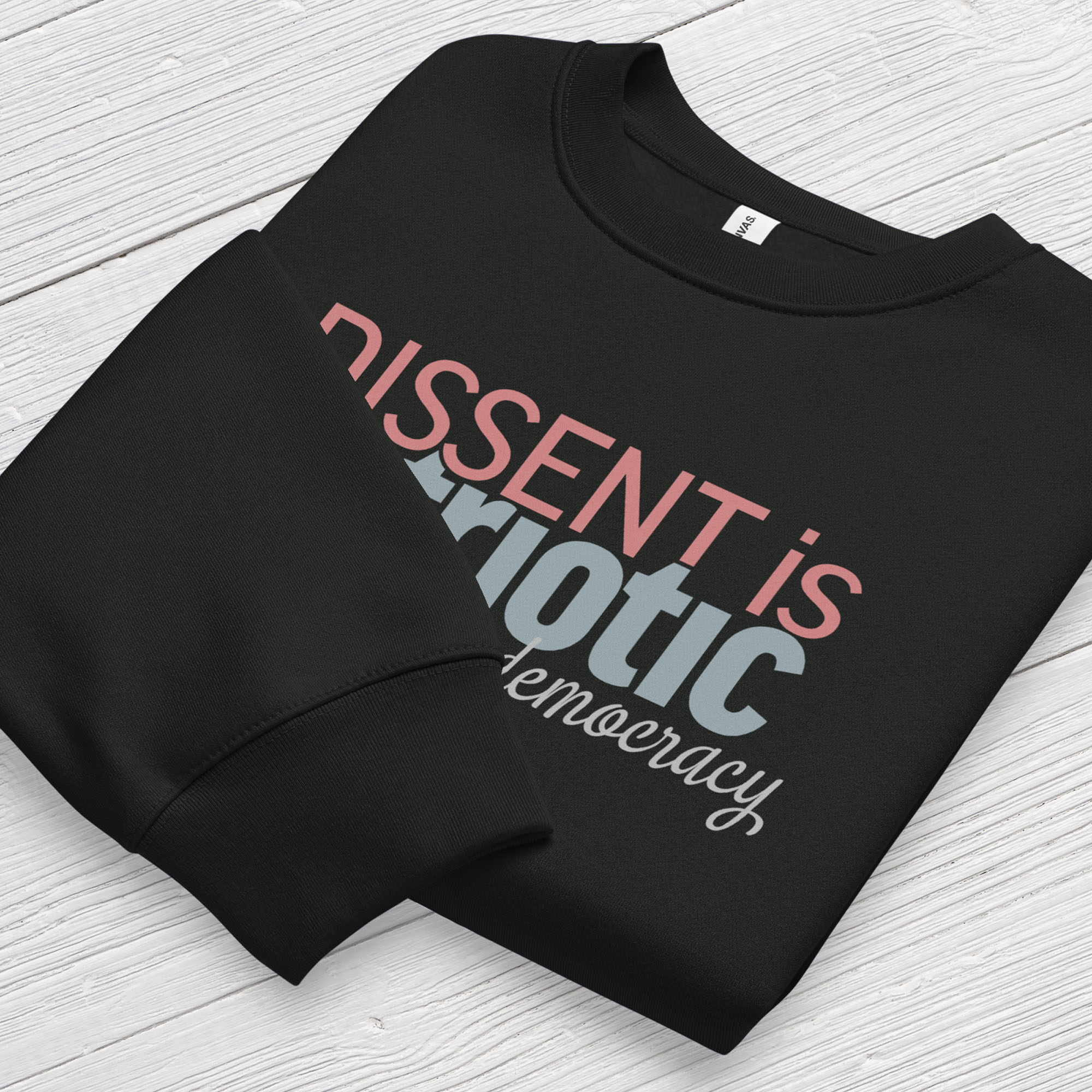 Folded black sweatshirt with bold stylized red and blue text that reads "dissent is patriotic" with script text below that reads "protest our democracy".