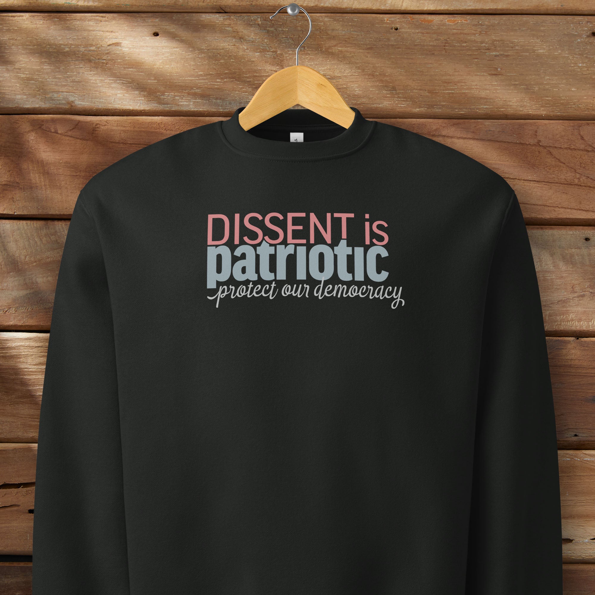 Black sweatshirt with bold stylized red and blue text that reads "dissent is patriotic" with script text below that reads "protest our democracy", hanging on a hanger.