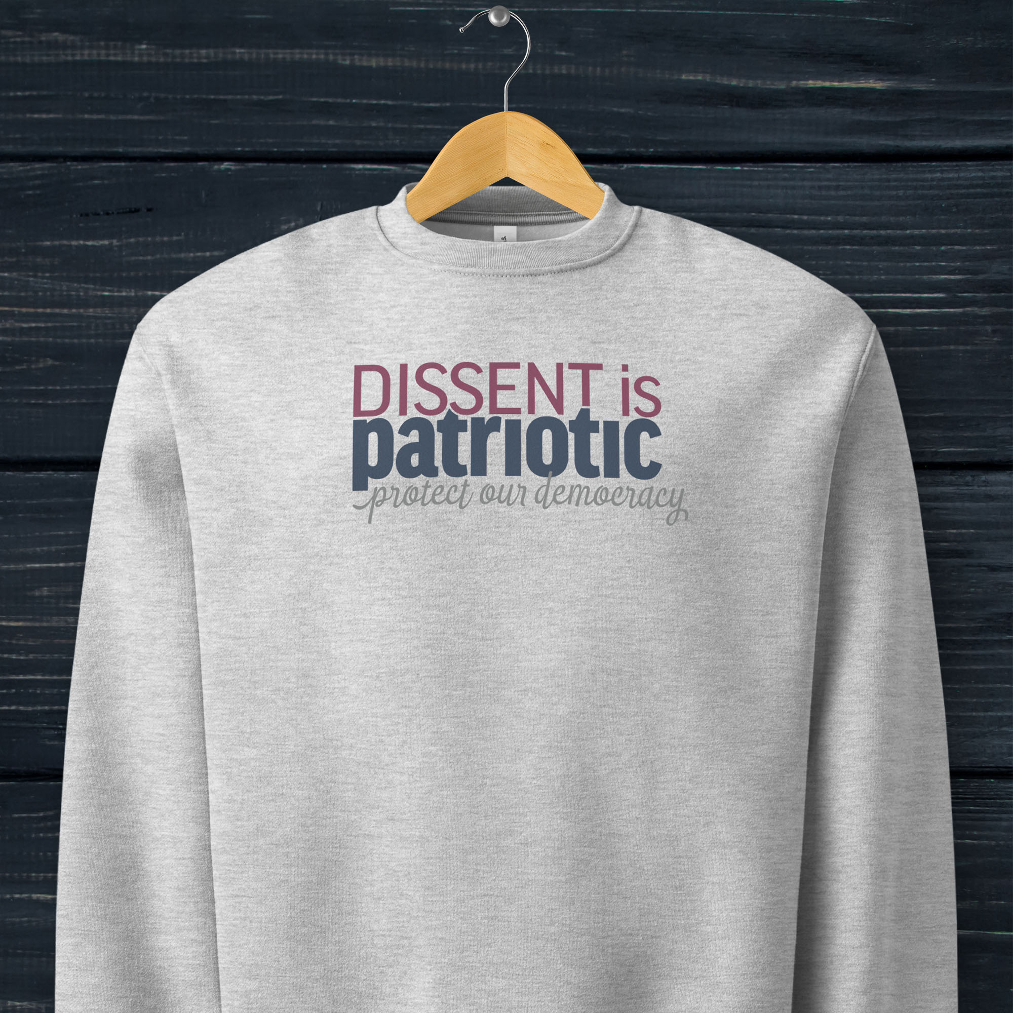 Athletic heather grey sweatshirt with bold stylized red and blue text that reads "dissent is patriotic" with script text below that reads "protest our democracy", hanging on a hanger.