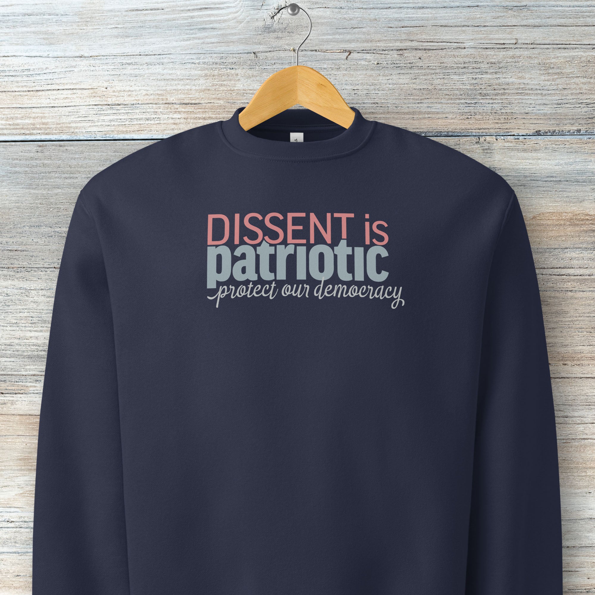 Navy sweatshirt with bold stylized red and blue text that reads "dissent is patriotic" with script text below that reads "protest our democracy", hanging on a hanger.