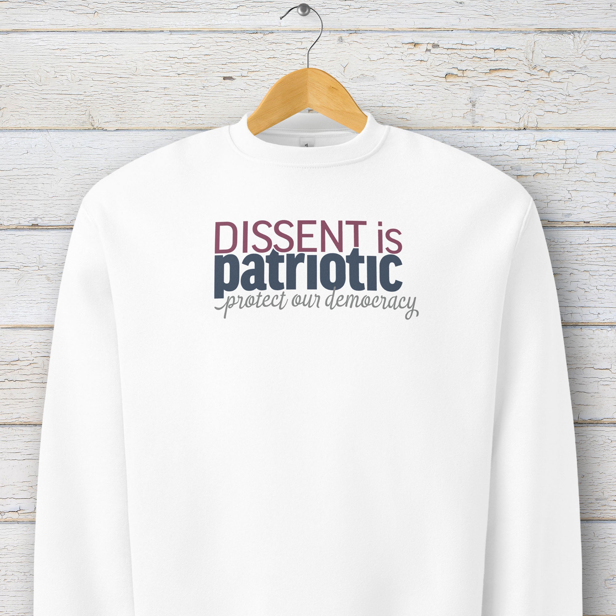 White sweatshirt with bold stylized red and blue text that reads "dissent is patriotic" with script text below that reads "protest our democracy", hanging on a hanger.