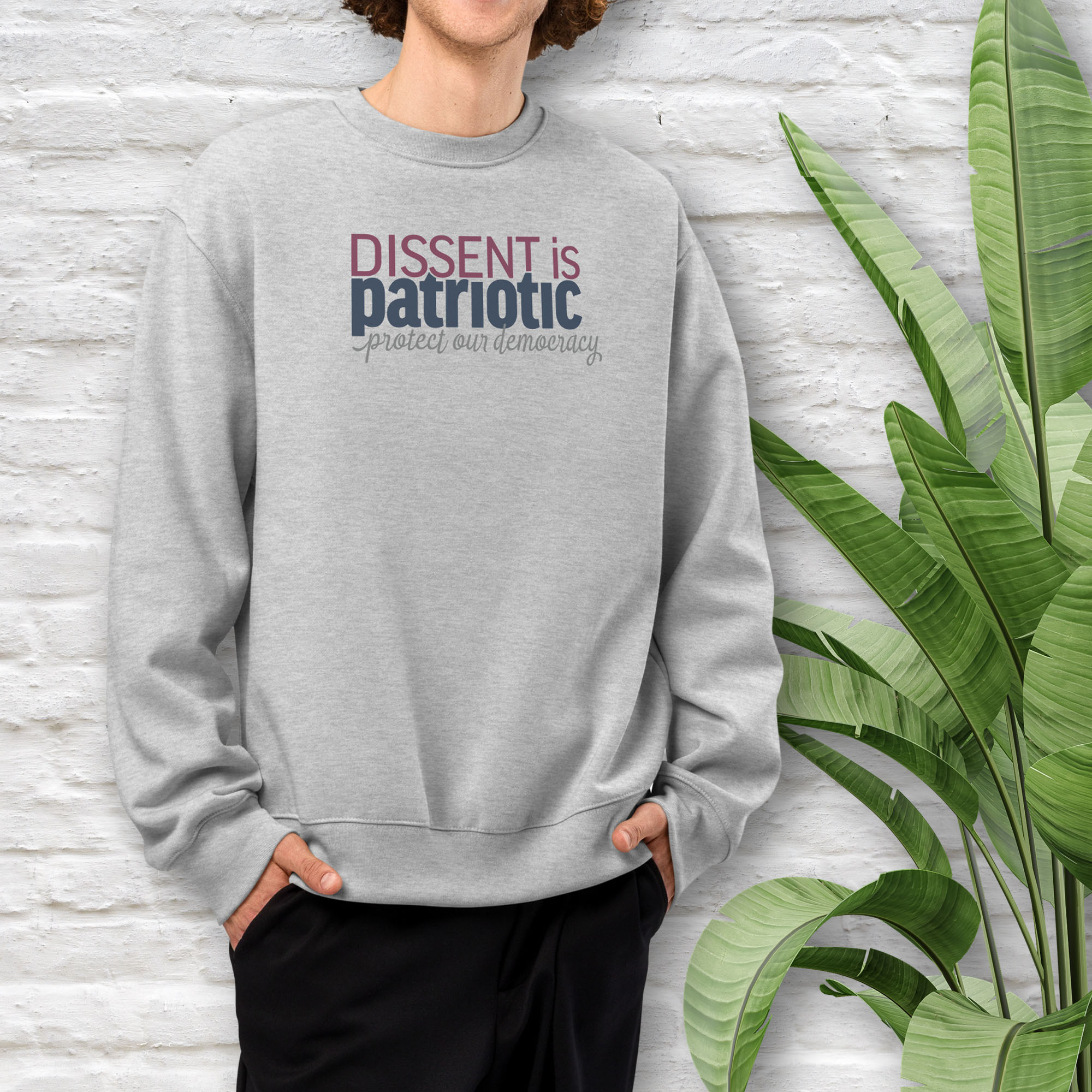 Male model wearing athletic heather grey sweatshirt with bold stylized red and blue text that reads "dissent is patriotic" with script text below that reads "protest our democracy".