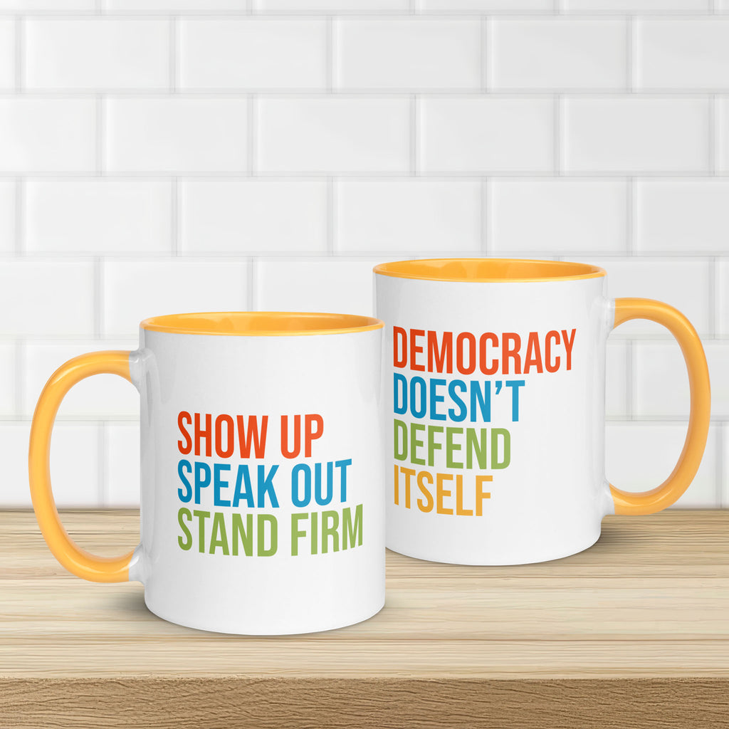 Two coffee mugs with bright gold handle and inside with text on one side that reads "show up, speak out, stand firm." The other side reads "democracy doesn't defend itself."