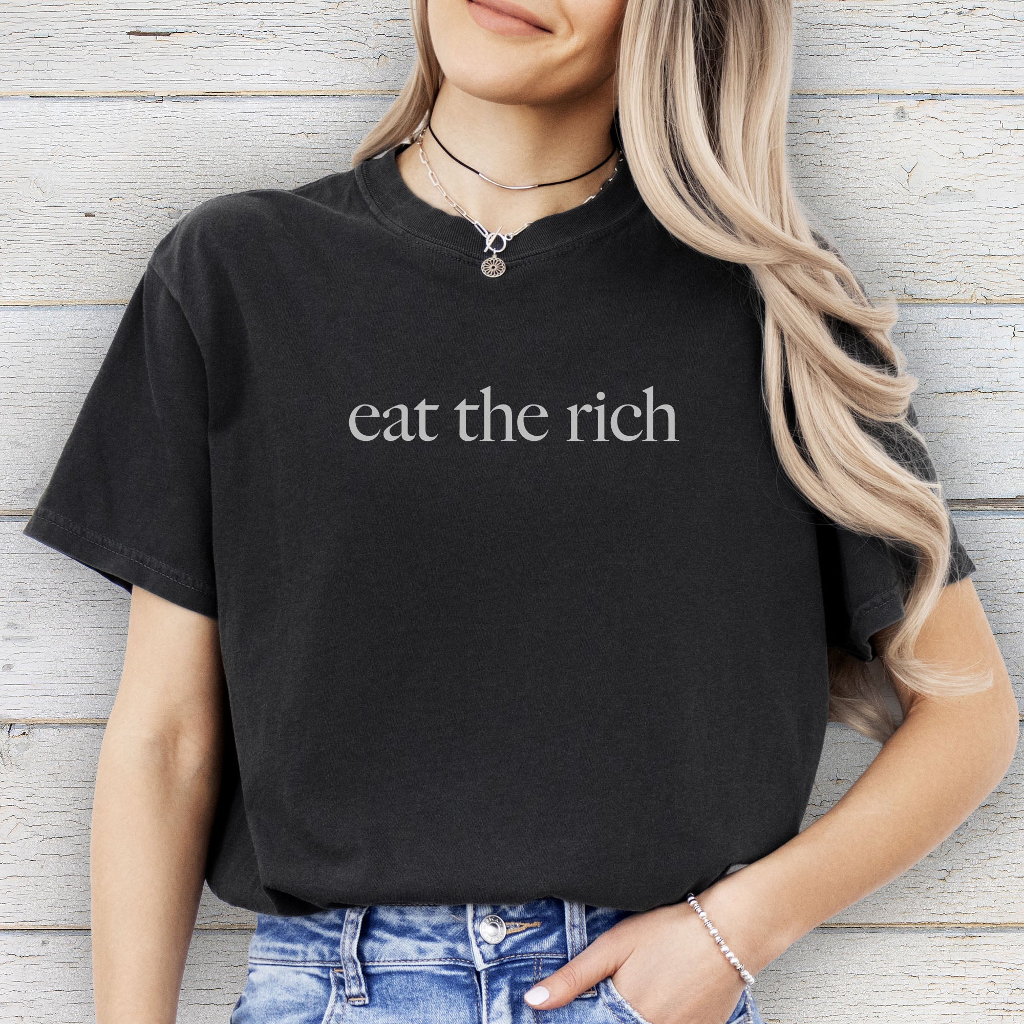 Female model wearing black t-shirt with minimalist light grey text reading “eat the rich.