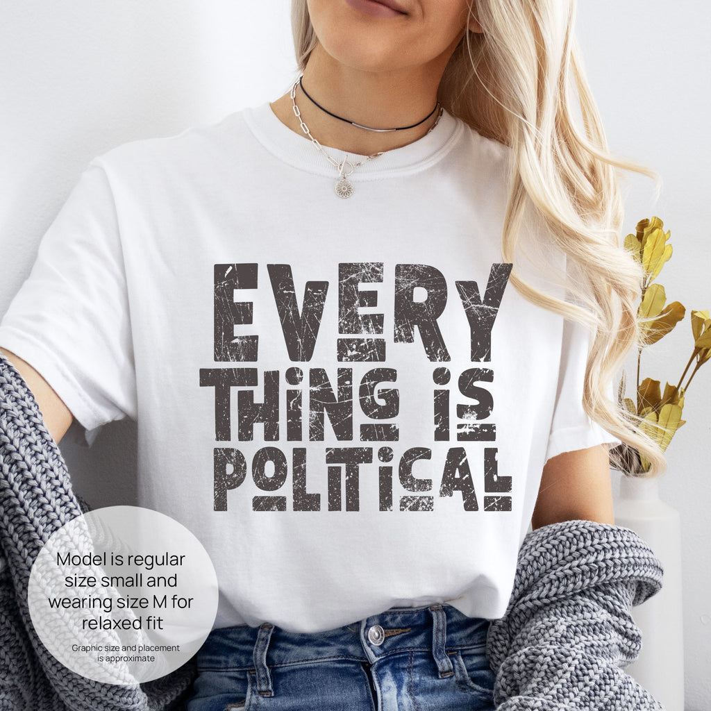 Female model wearing white t-shirt featuring distressed block typography reading "Everything Is Political" in a bold minimalist design. Includes size and fit information for the model who is regular size small, and wearing a size M shirt for a relaxed fit.
