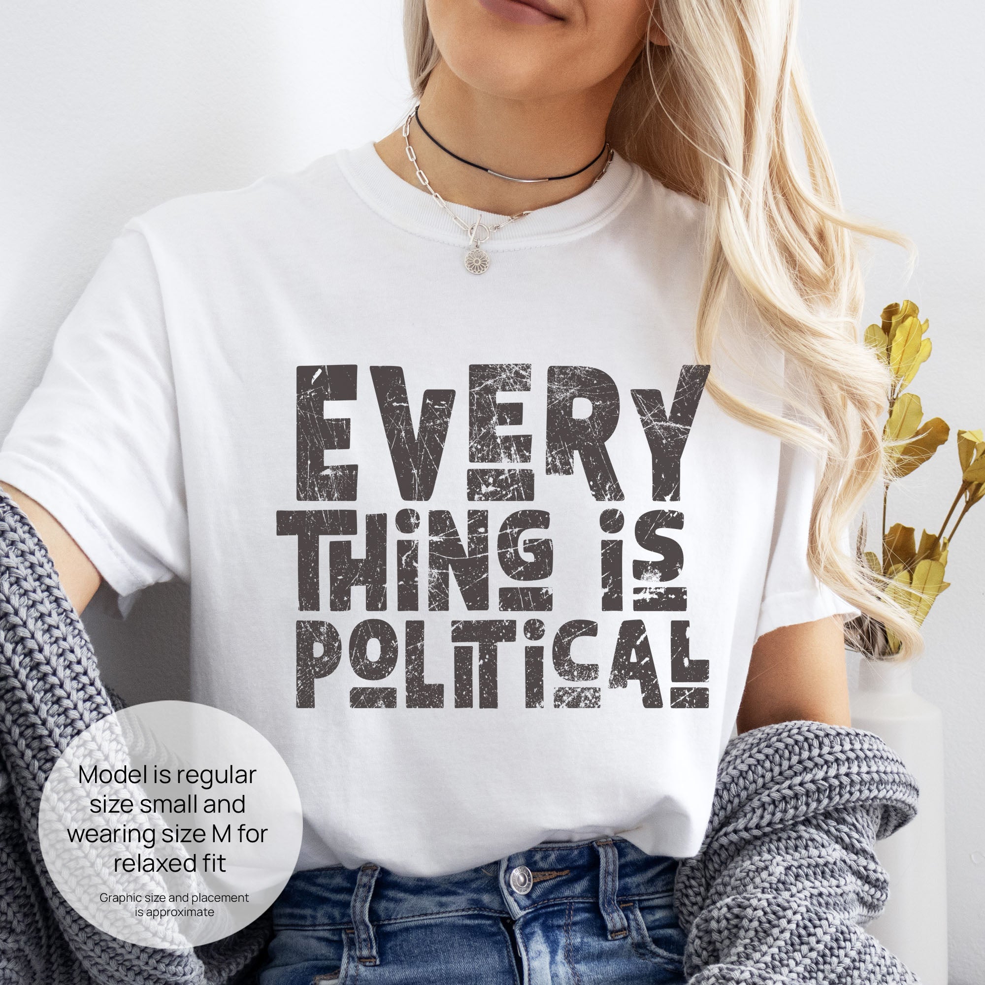 Female model wearing white t-shirt featuring distressed block typography reading "Everything Is Political" in a bold minimalist design. Includes size and fit information for the model who is regular size small, and wearing a size M shirt for a relaxed fit.
