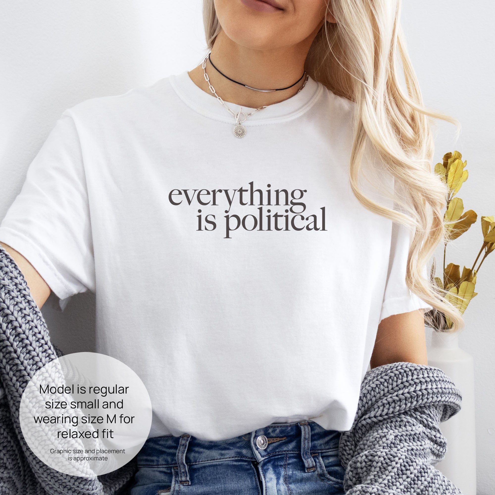 Female model wearing white unisex t-shirt with minimalist serif gray text reading “everything is political." Includes size and fit information for the model who is regular size small, and wearing a size M shirt for a relaxed fit.
