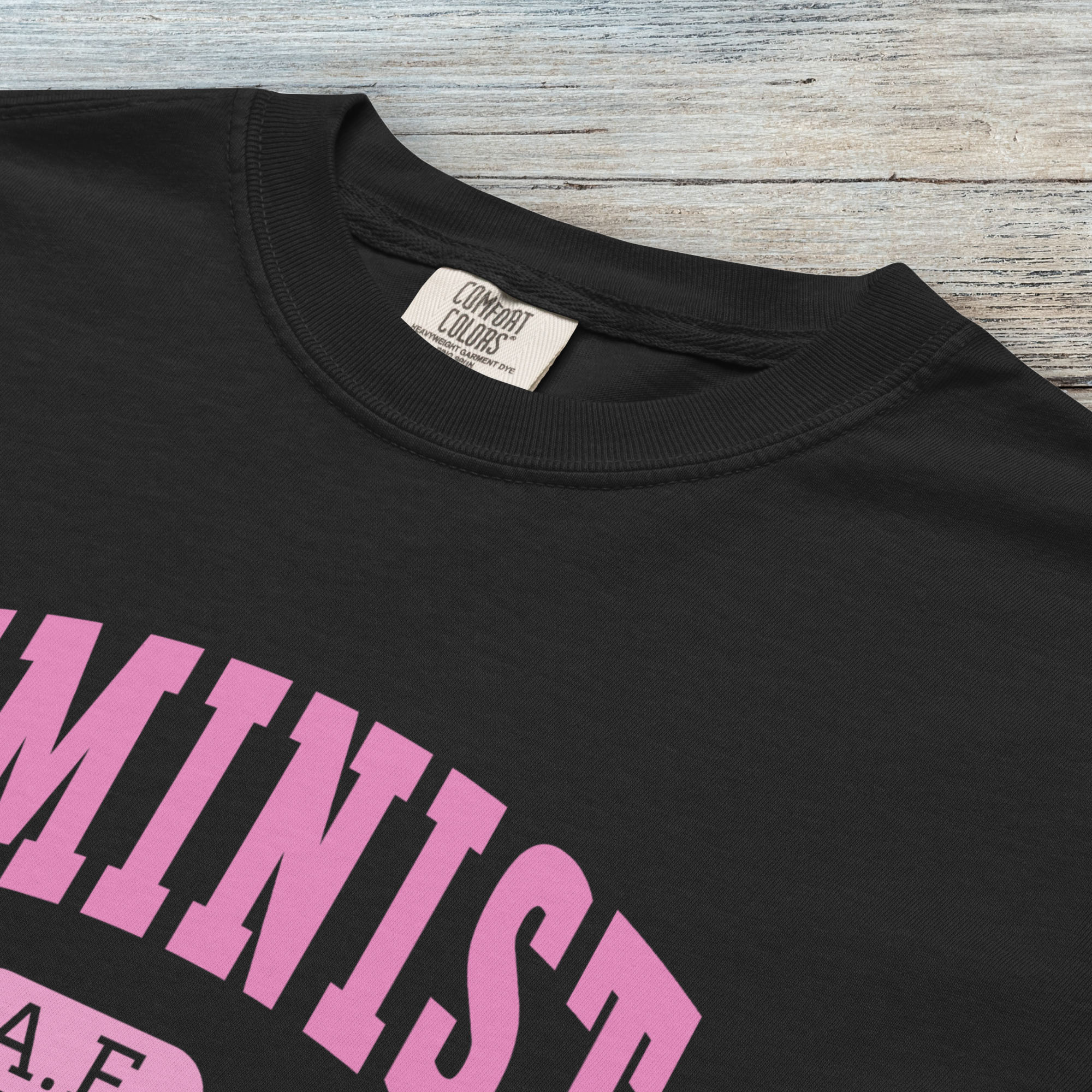 Closeup detail of black Comfort Colors tee with pink text that reads “Feminist A.F.” in a collegiate style.