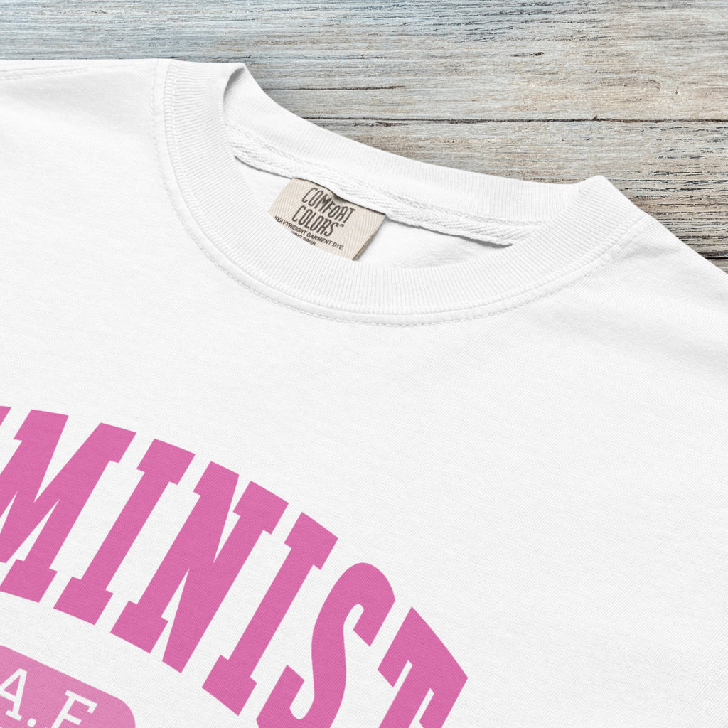 Closeup detail of white Comfort Colors tee with pink text that reads “Feminist A.F.” in a collegiate style.