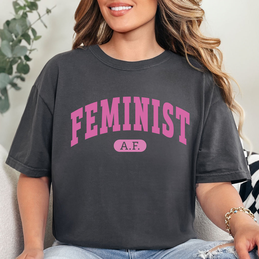 Female model wearing graphite Comfort Colors tee with pink text that reads “Feminist A.F.” in a collegiate style.