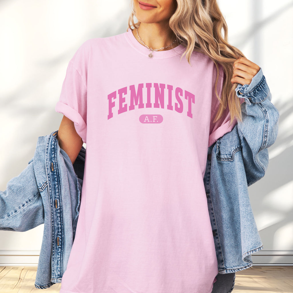 Female model wearing blossom Comfort Colors tee with pink text that reads “Feminist A.F.” in a collegiate style.