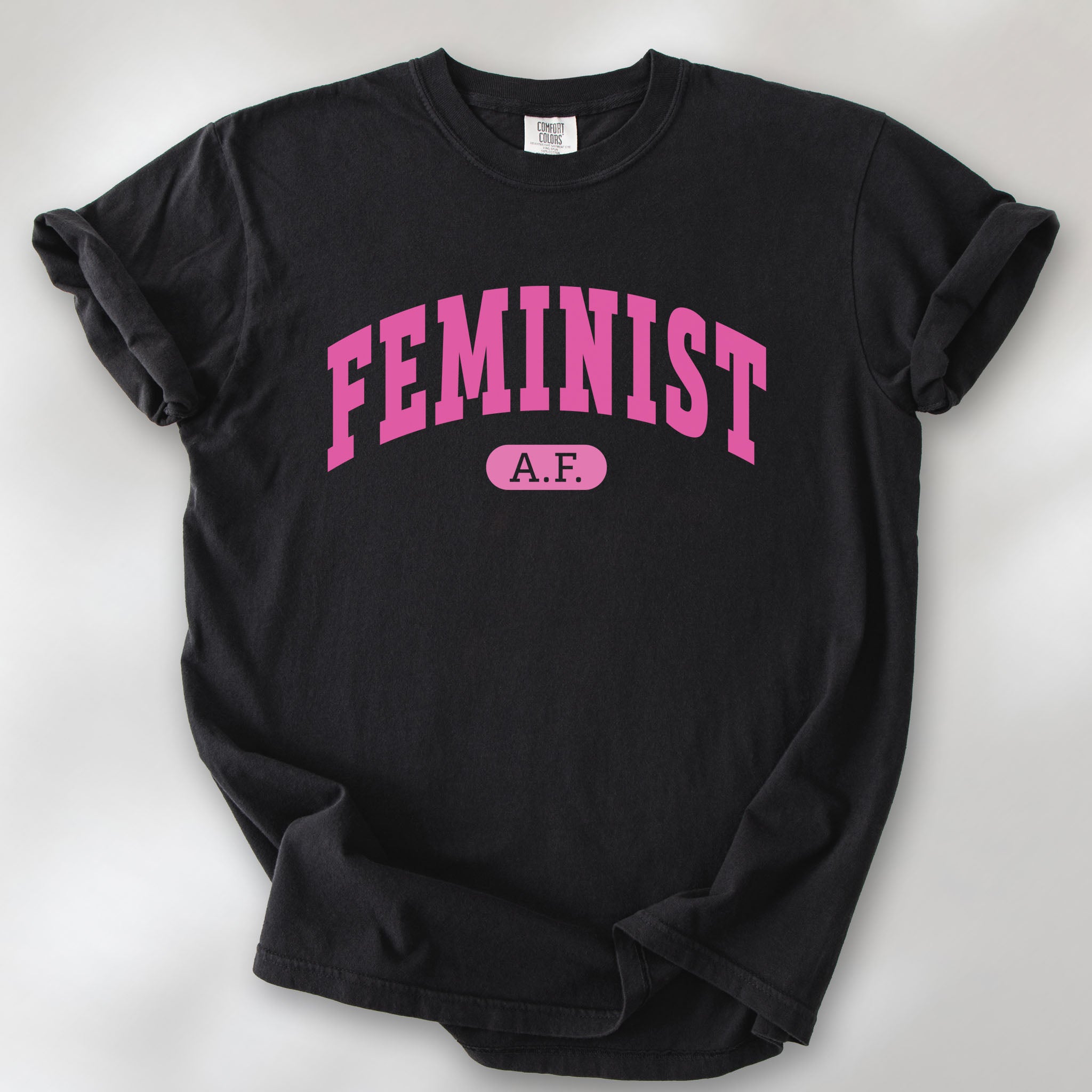 Black Comfort Colors tee with pink text that reads “Feminist A.F.” in a collegiate style.