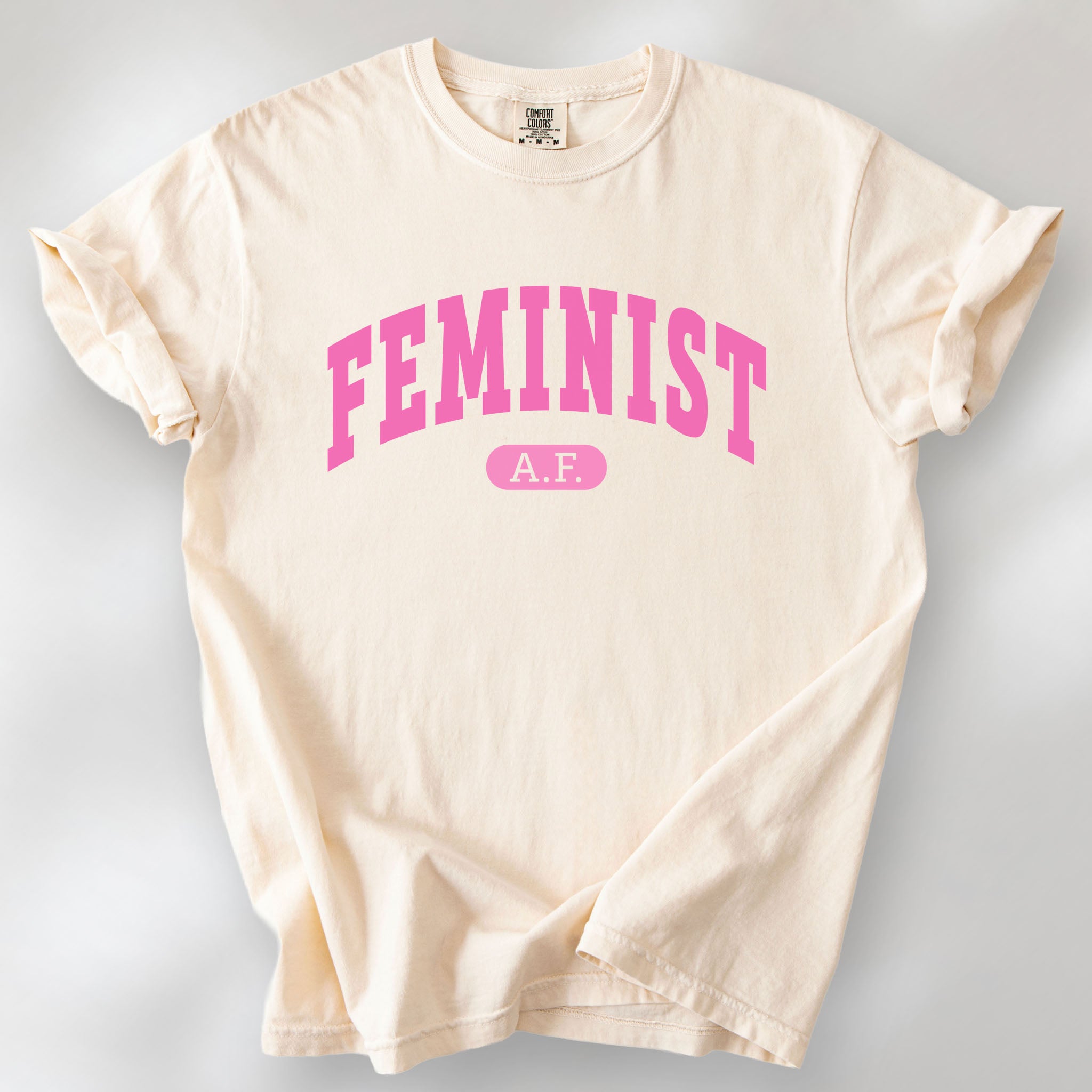 Ivory Comfort Colors tee with pink text that reads “Feminist A.F.” in a collegiate style.