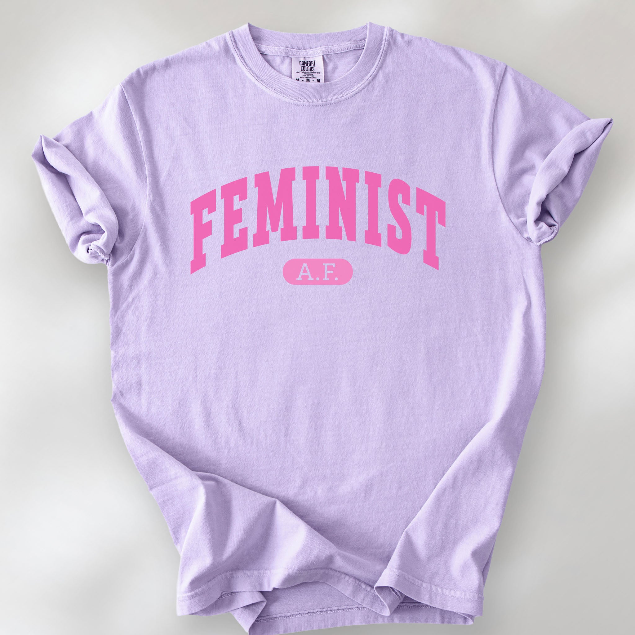 Orchid Comfort Colors tee with pink text that reads “Feminist A.F.” in a collegiate style.